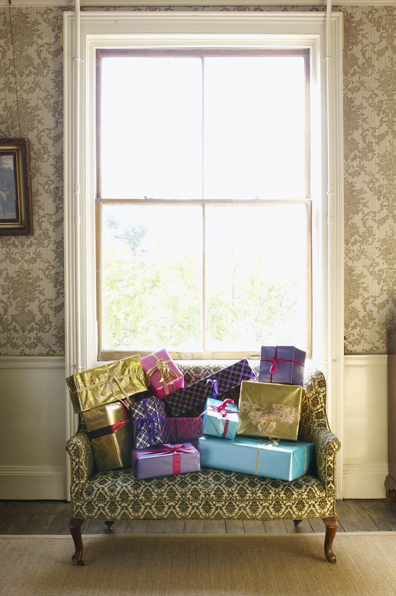 A vintage-style sofa with a floral pattern, filled with colorful wrapped presents, in front of a large window with bright sunlight.