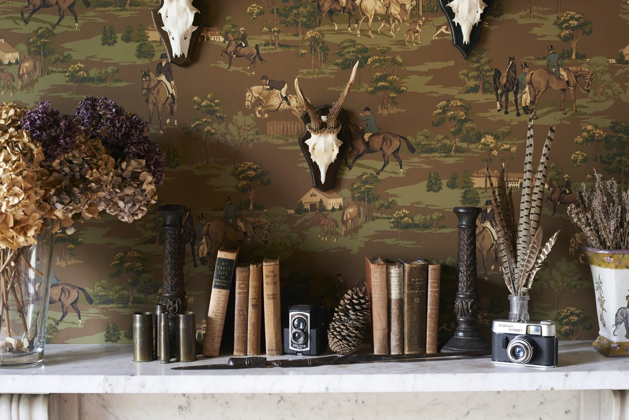 Decorative shelf with dried flowers, antique books, a pinecone, a vintage camera, and a small speaker, set against a wallpaper with a hunting scene featuring mounted hunters and animals.