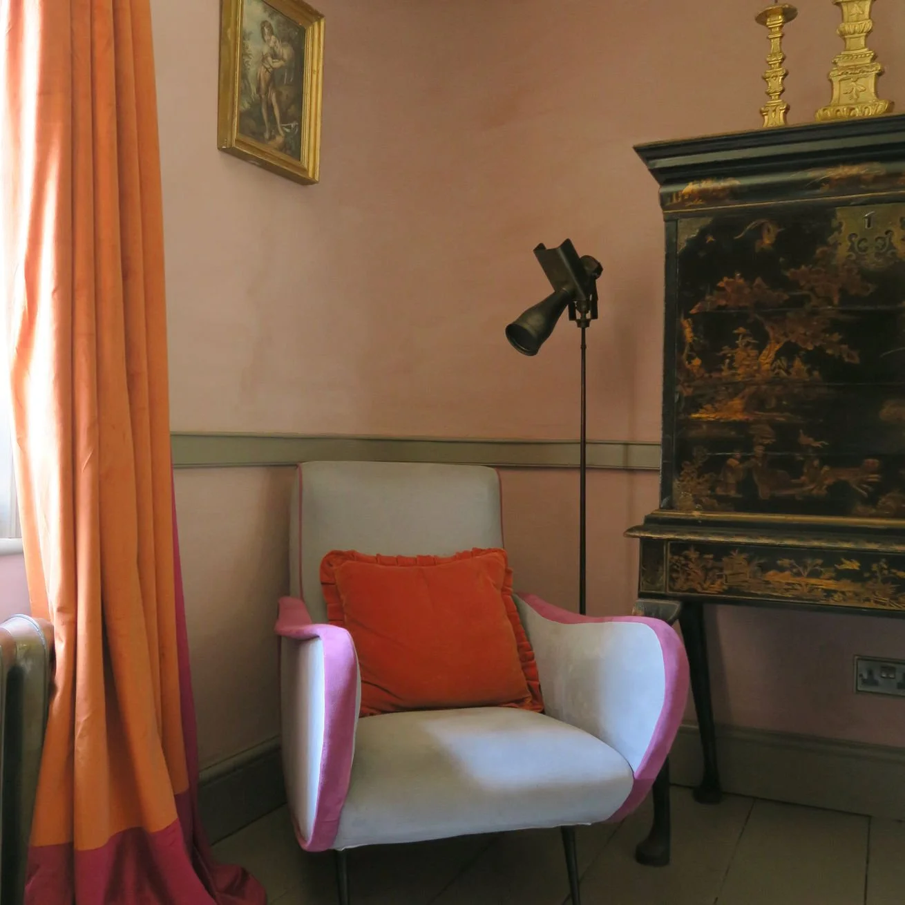 A room with a beige armchair with pink edges and an orange pillow, orange curtains on the left, a decorative dark green cabinet with gold details on the right, a floor lamp next to the armchair, and a framed painting on the wall above.
