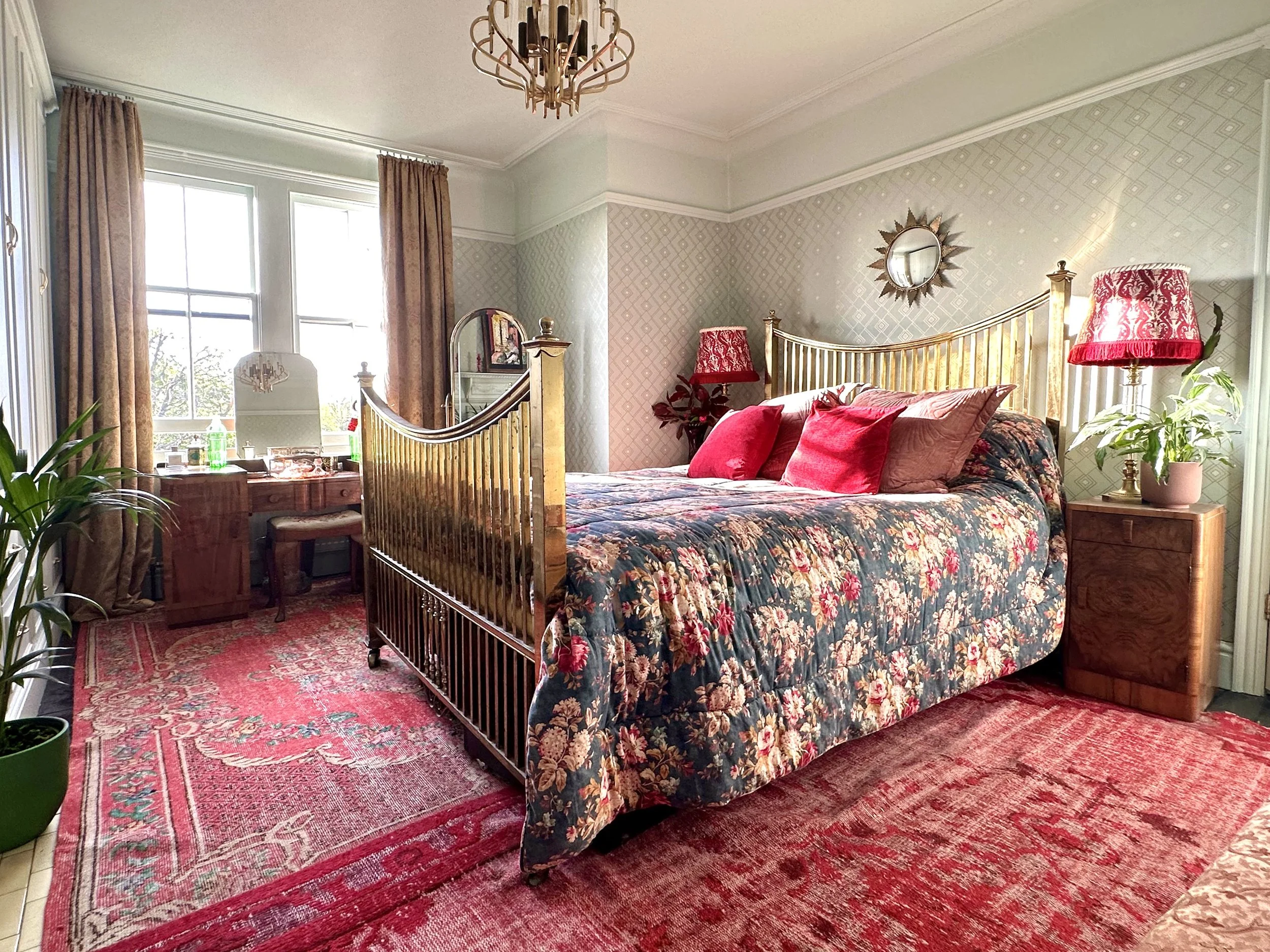 A bedroom with a large bed featuring a floral patterned quilt and multiple red and beige pillows. There are two wooden nightstands with red lamps on each side of the bed. A gold-colored headboard is present. A round mirror hangs on the wall above the