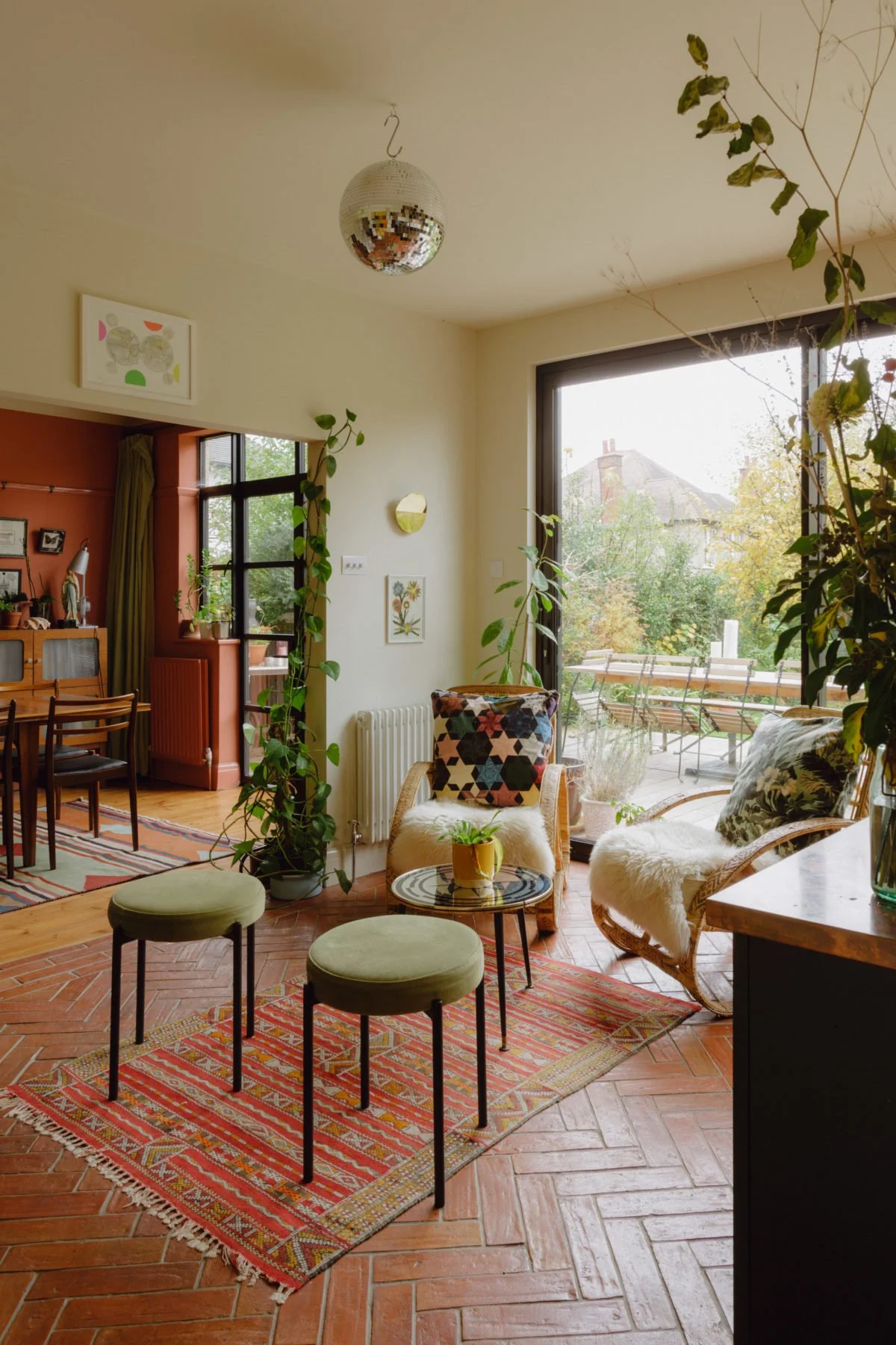 A cozy living room with two green cushioned stools, a small round table with a potted plant, and two wicker chairs with fur throws and patterned cushions, set on a colorful rug near large windows showing a balcony and outdoors.