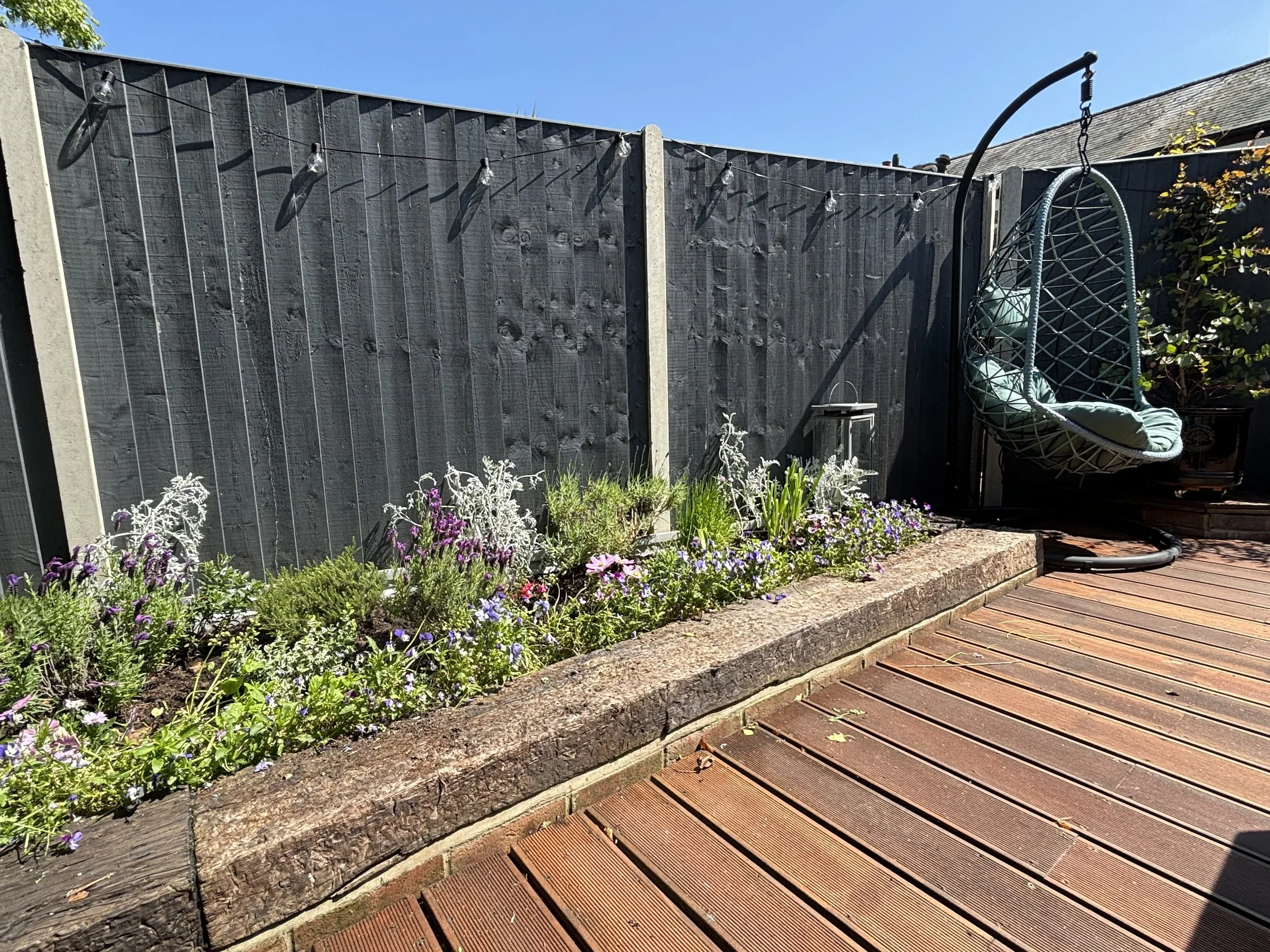 A backyard with a wooden deck, a flower bed with purple, white, and green plants, black painted wooden fence with lights, and a hanging egg-shaped swing chair with teal cushions.