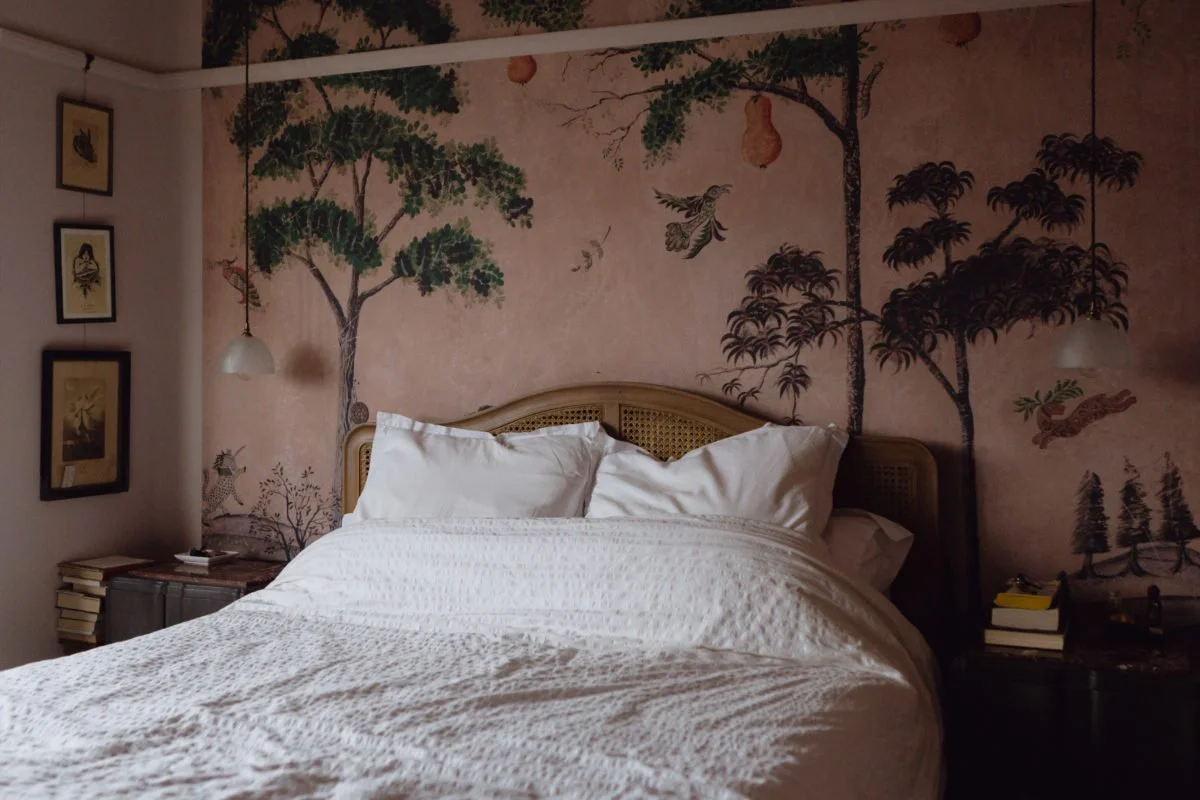 A bedroom with a mural of trees, birds, and a tiger on the wall behind the bed, which has white bedding and pillows, nightstands with lamps, books, and decorative items.