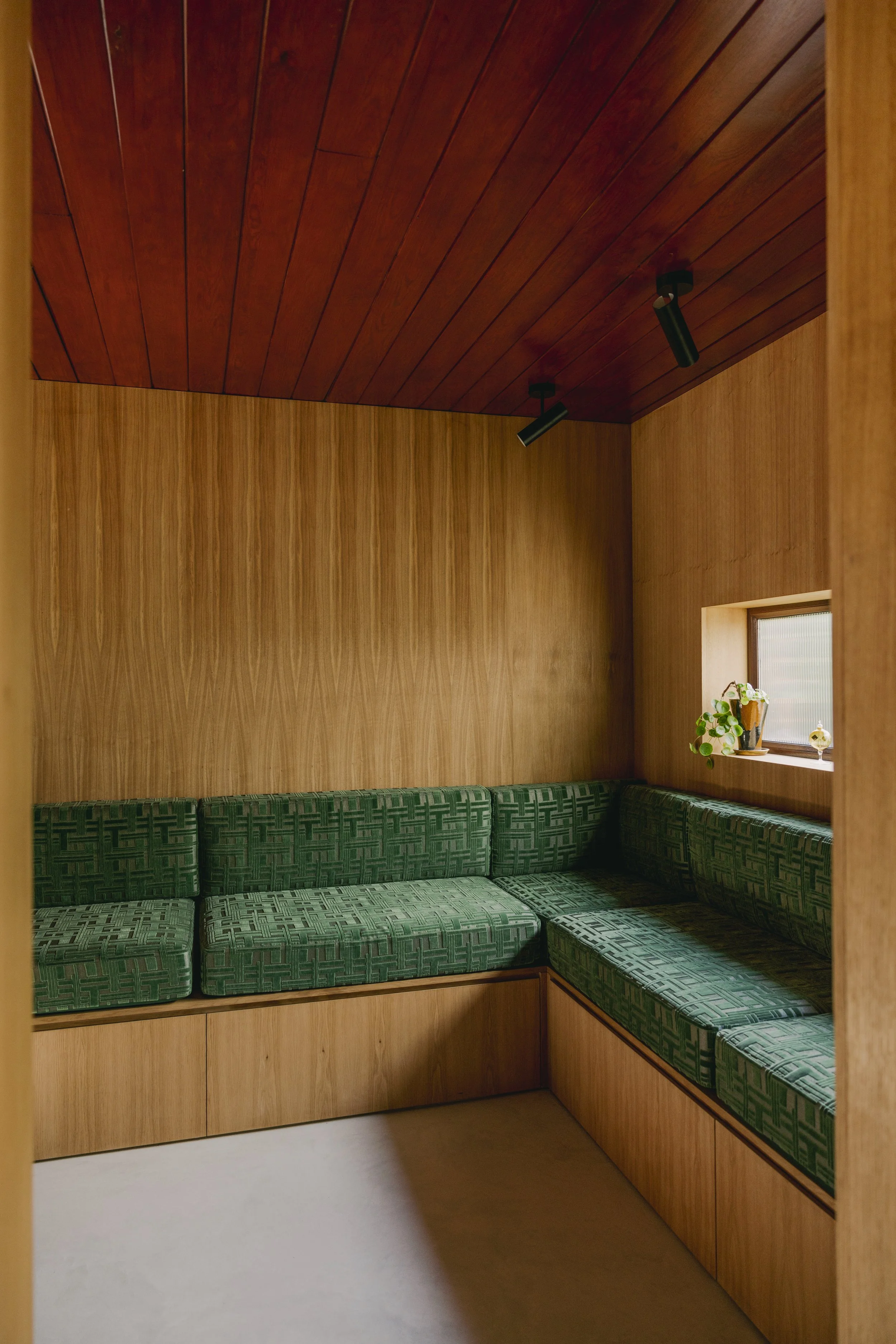 A corner with green patterned cushioned seating built into wooden walls and a wooden ceiling, featuring a small window with potted plants on the windowsill.