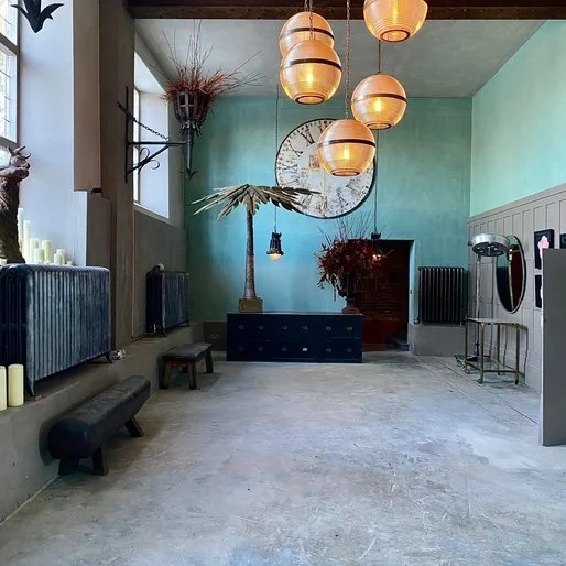 Interior of a modern salon or spa with hanging pendant lights, a large wall clock, a tall palm plant, and various furniture including benches and station stations.