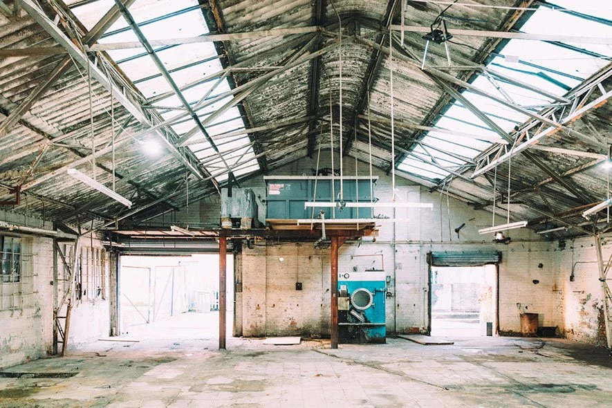 Empty, dilapidated industrial warehouse with a high, broken, metal roof and concrete walls, featuring laundry machines in the center and large open doorways.