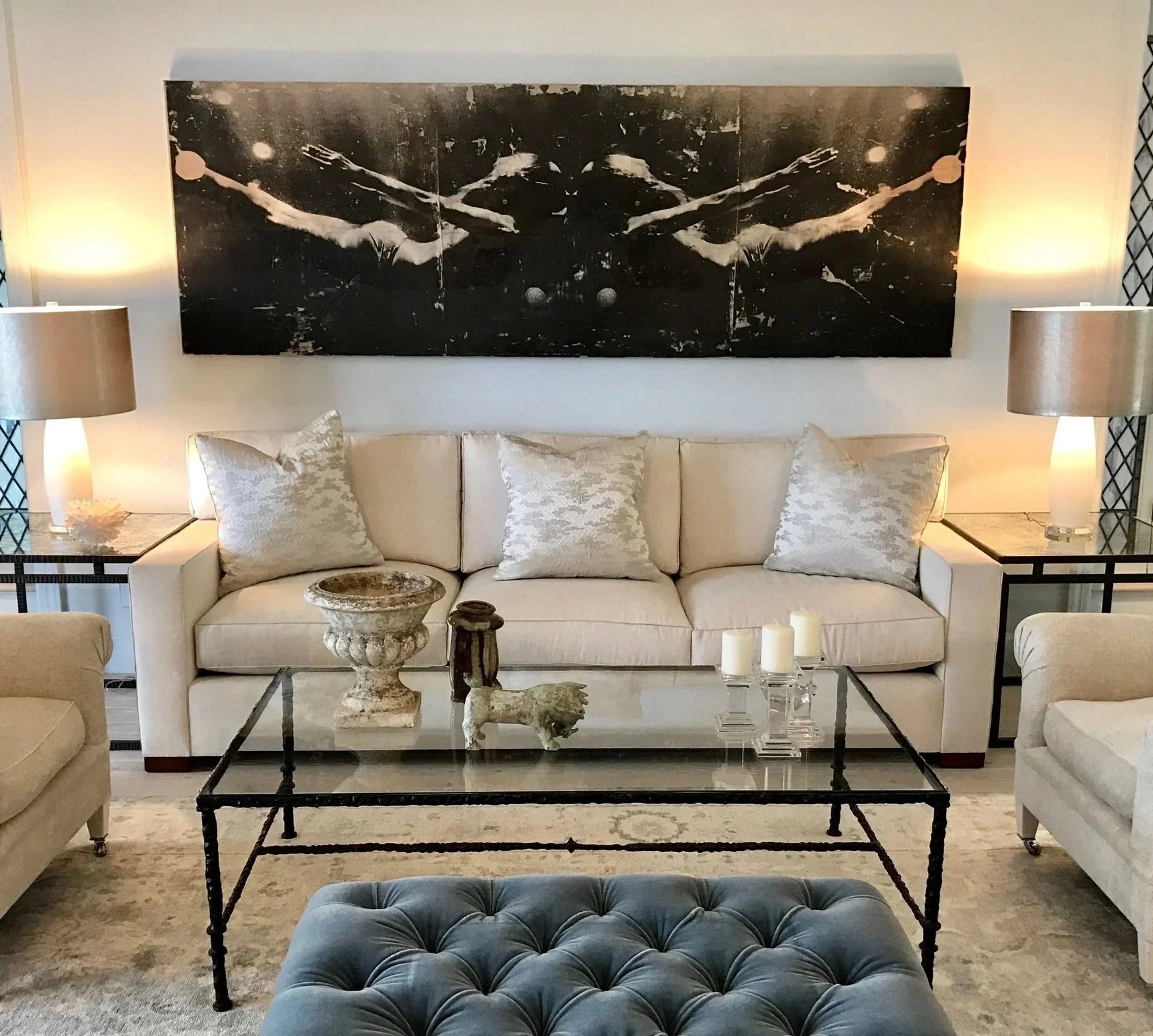 A beige sofa with four pillows, flanked by two side tables with matching lamps. A black and white artwork hangs on the wall above the sofa. In front, a glass coffee table with decorative items and four white candles. Two beige armchairs on each side,