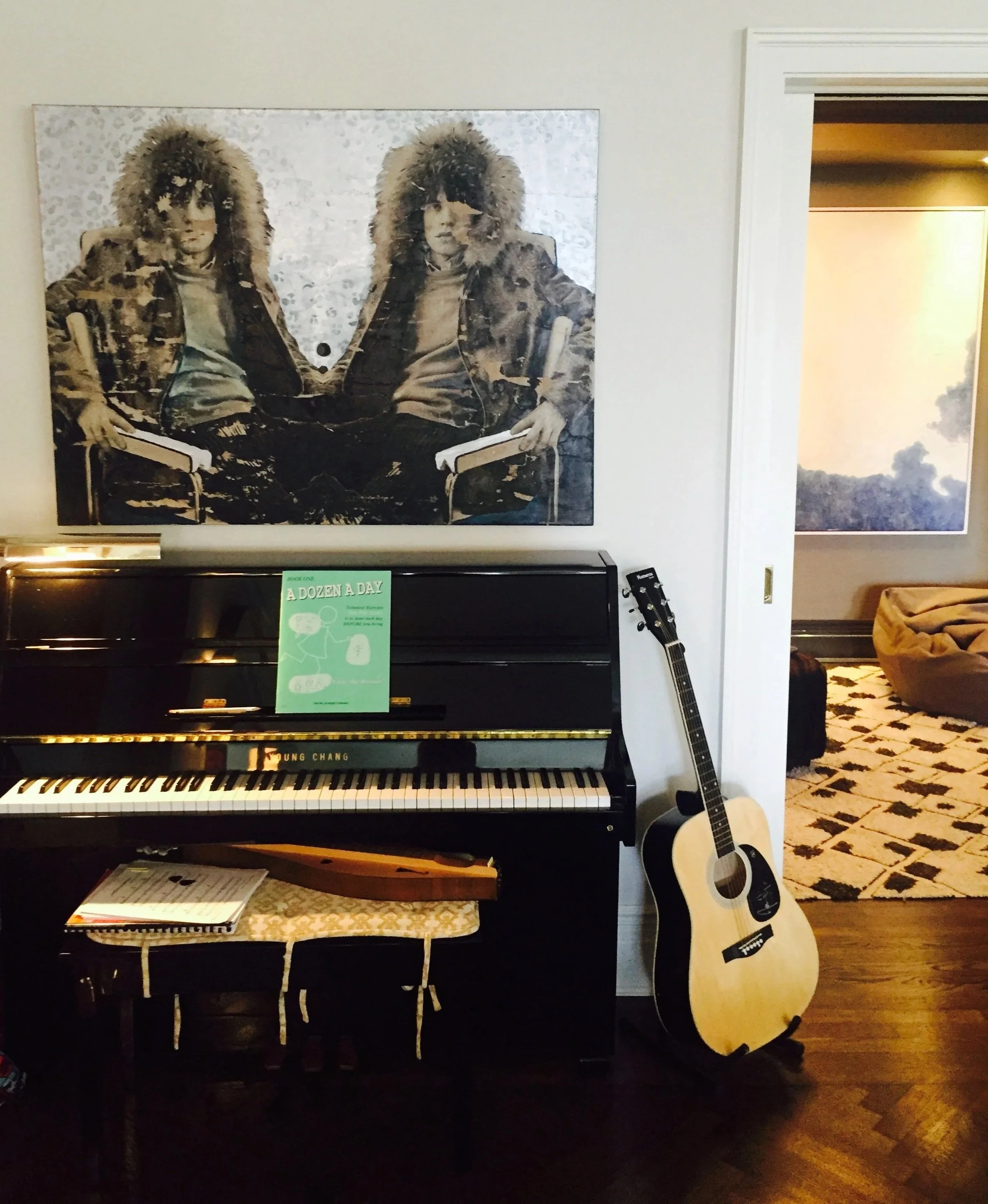 A black upright piano with a music book open on top, next to a guitar leaning against the wall, in a room with a large framed picture of two people in fur-lined hoods on the wall. There is a patterned area rug visible in the adjoining room.