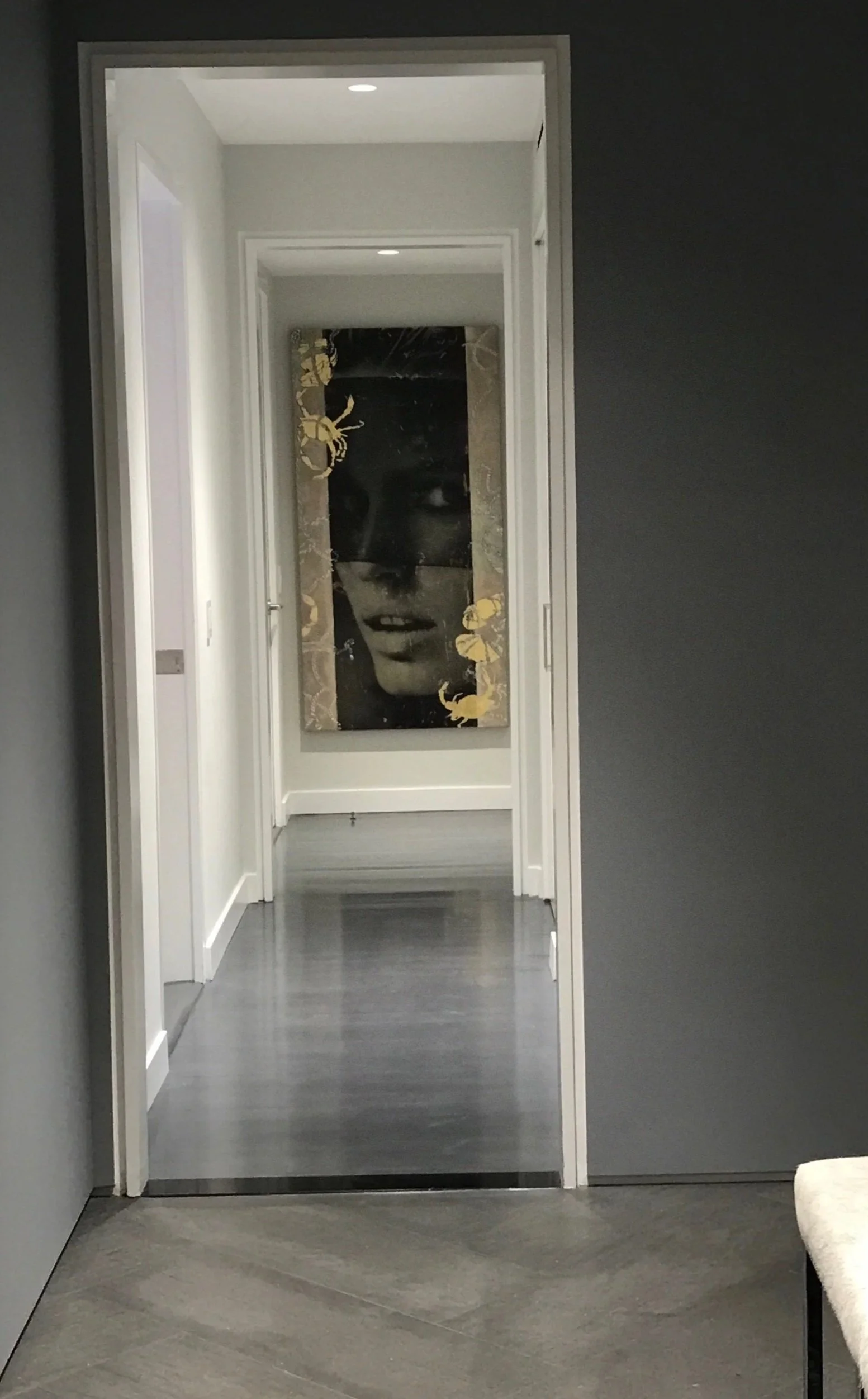 View of a modern hallway with gray walls and flooring, leading to a room with an artistic black and white portrait of a woman's face, surrounded by decorative golden elements.