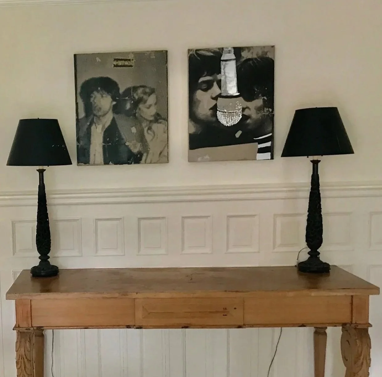 A wooden side table with two black lamps on top, against a wainscoted white wall with black-and-white framed photos of people. To the left, part of a dining table with a vase of white flowers and some magazines, and a chandelier with antler-like bran