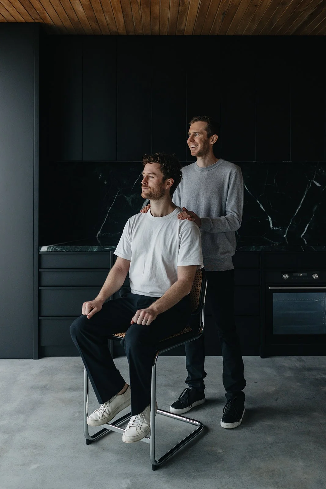 Two men in a modern kitchen, one sitting on a chair and the other standing behind him with hands on his shoulders, both smiling and appearing relaxed.