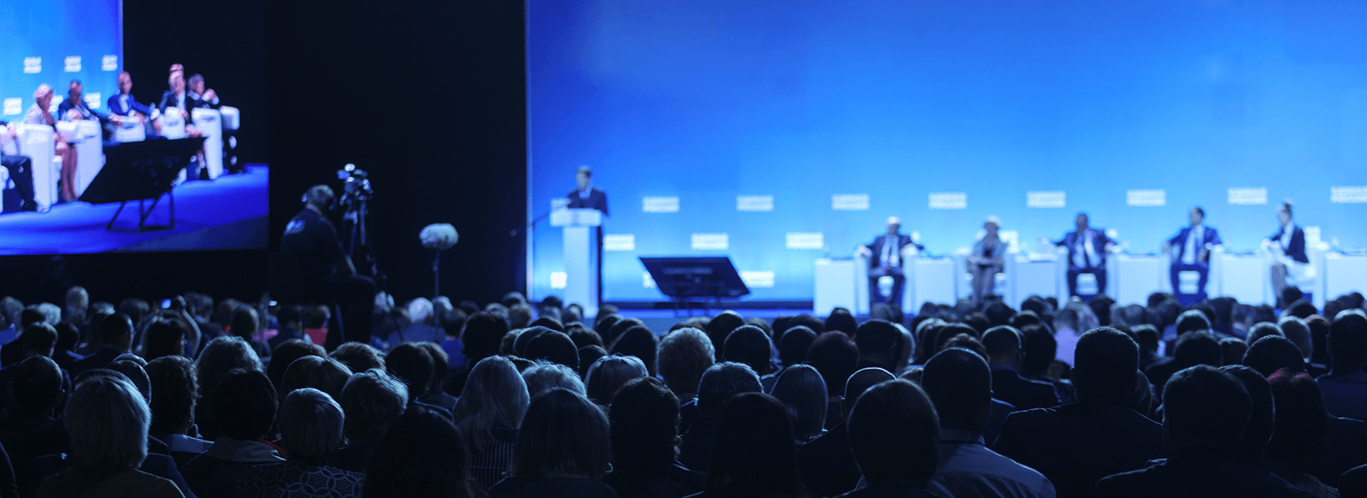 Audience watching a panel discussion and keynote presentation at a large business event in a blue-lit conference venue