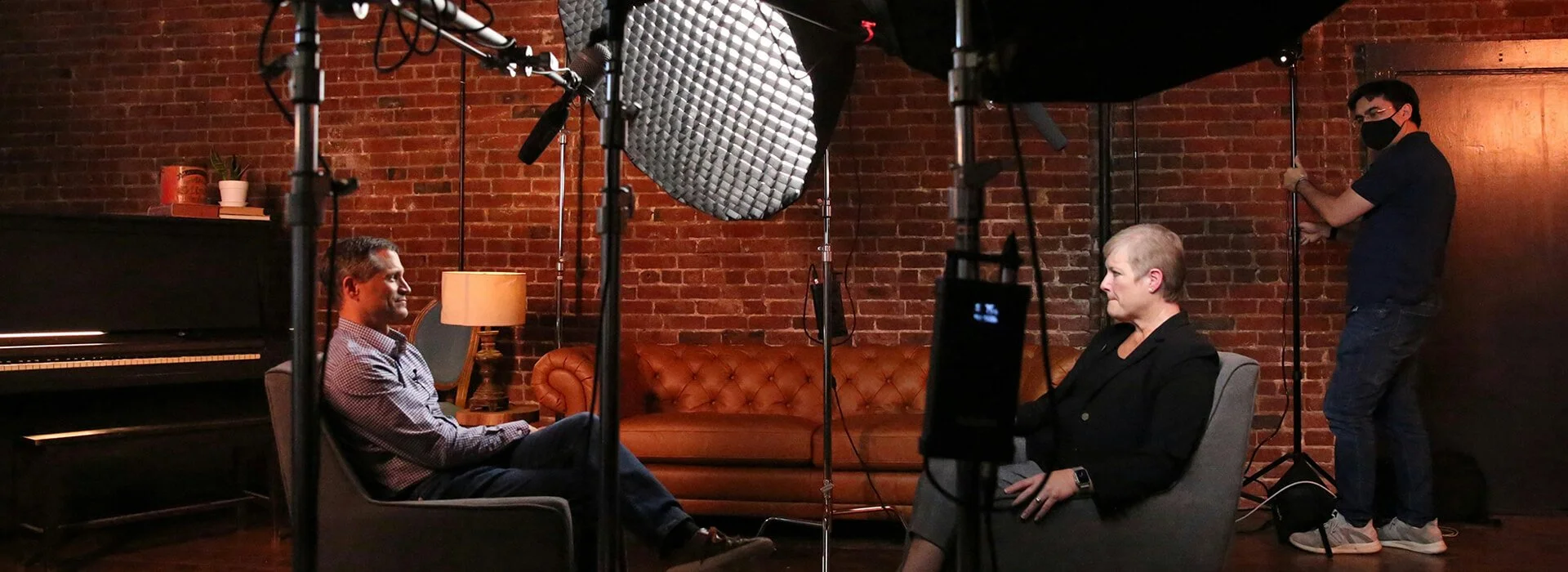Behind-the-scenes view of a business video interview setup with two people seated opposite each other, studio lights, boom microphones, and a crew member adjusting lighting in a brick-walled studio.