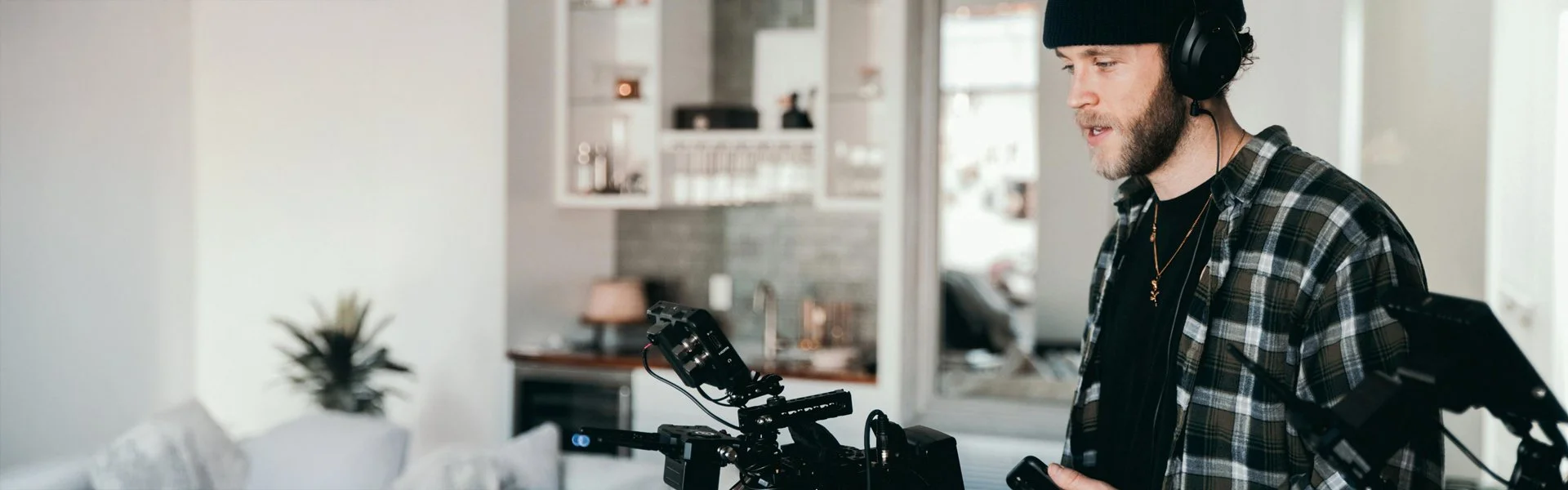 Man wearing headphones and a plaid shirt operating a camera in a modern living room with plants and furniture