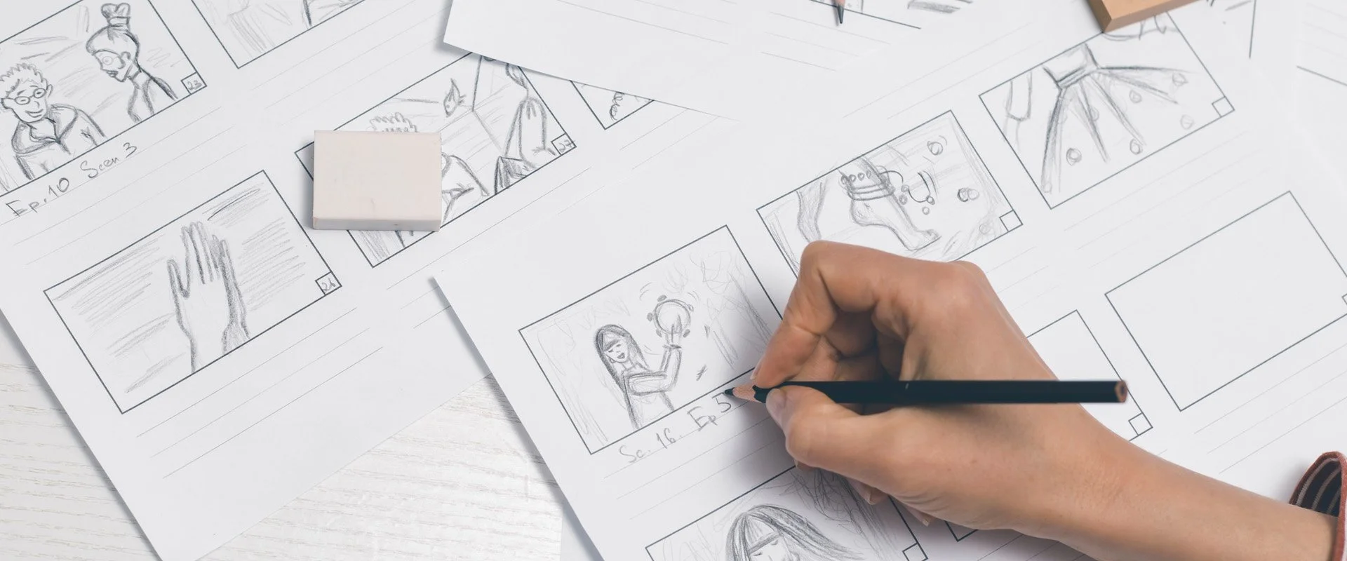Hand sketching a storyboard with pencil on paper, featuring detailed frames of characters and scenes, surrounded by eraser and additional storyboard sheets, ideal for video production planning.
