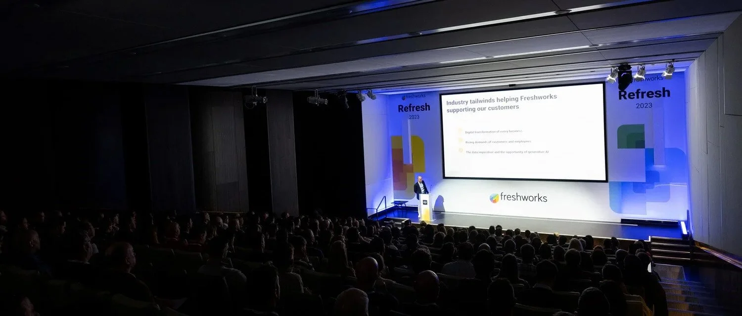 Speaker presenting on stage at Freshworks Refresh 2023 conference in London, addressing a large seated audience with branding and slides visible