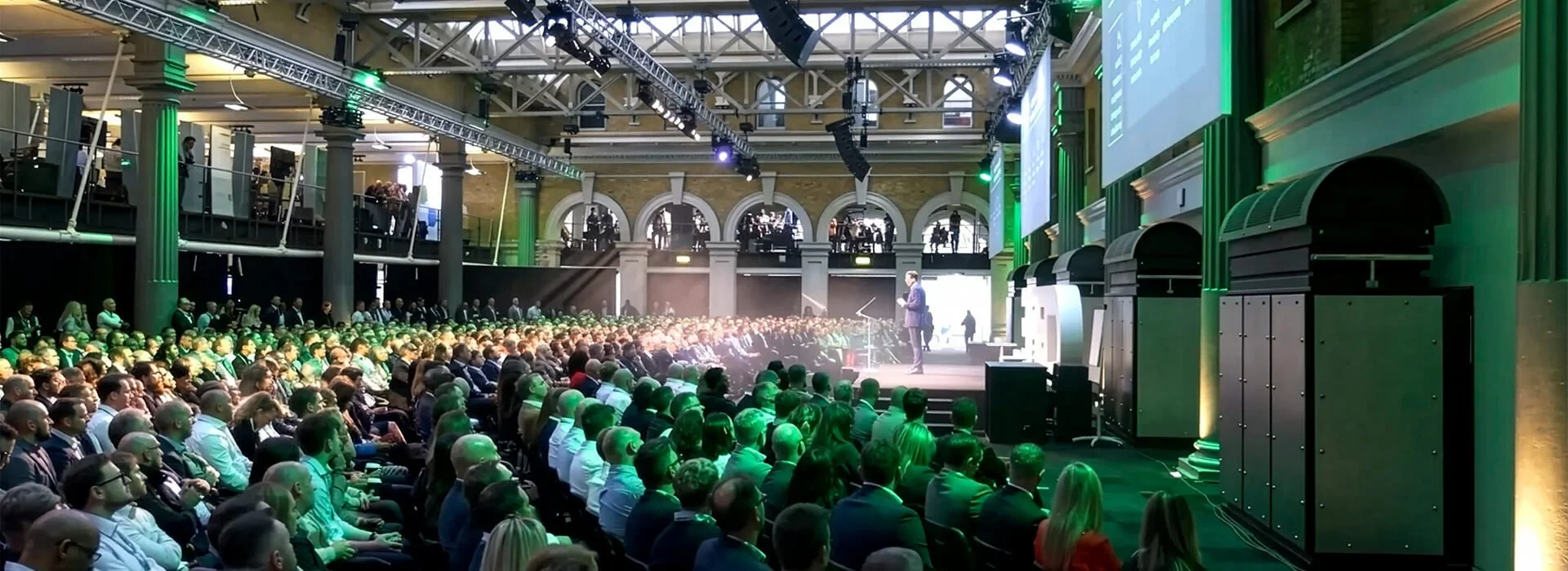 A large audience in business attire listens to a speaker in a spacious venue with a high, arched ceiling, green lighting, and a large screen. Tall columns and overhead lighting rigs are visible.