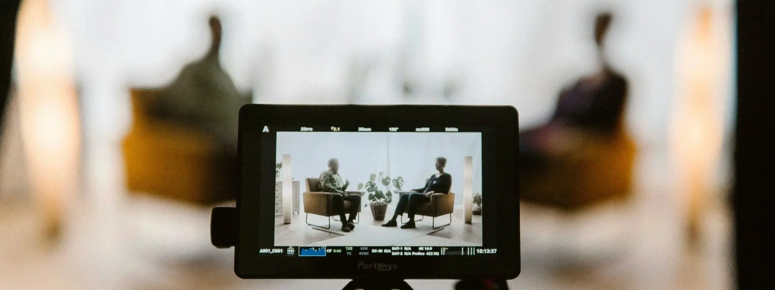 Camera monitor showing two people seated for an interview, with the filmed scene in focus on screen and the real set softly blurred in the background.