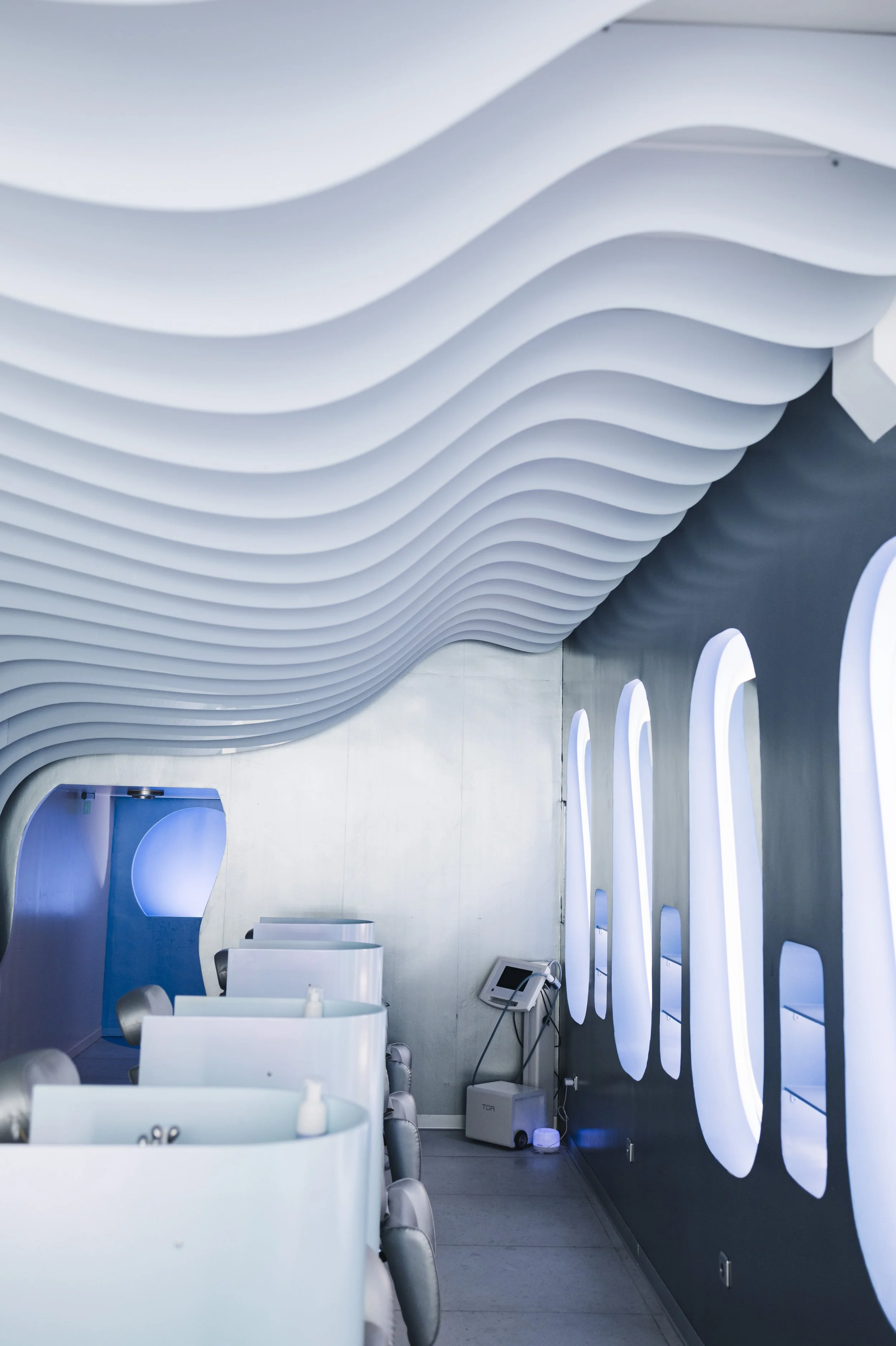 Modern interior with wavy white ceiling design, large oval-shaped windows on dark wall, and white seating area featuring tables and chairs.