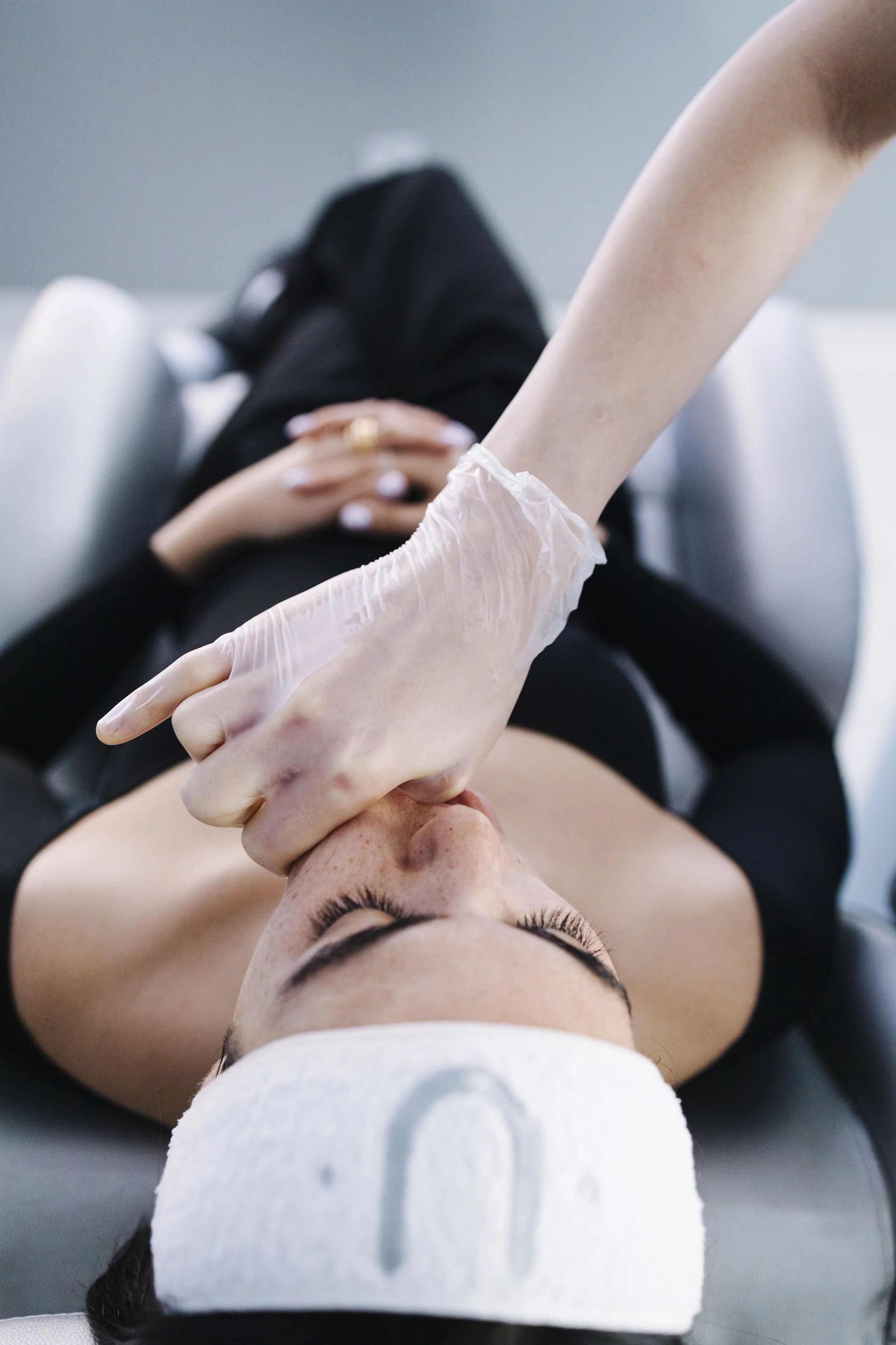 A woman receiving a facial treatment with a cotton pad on her face, lying on a treatment bed, with her eyes closed and wearing a white headband.