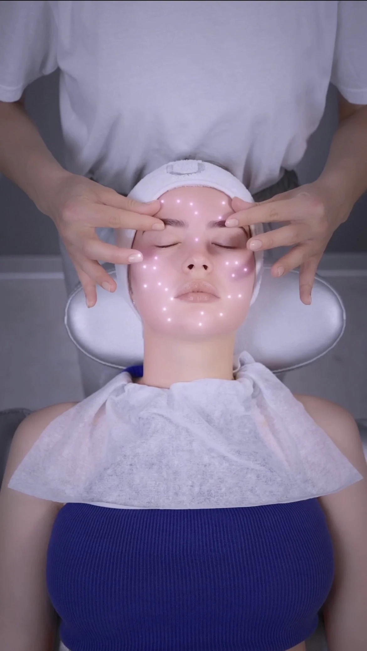 A woman receiving a facial treatment at a spa, lying on a bed with her eyes closed, wearing a blue top and a white towel; a professional applies skincare or massage to her face with both hands.