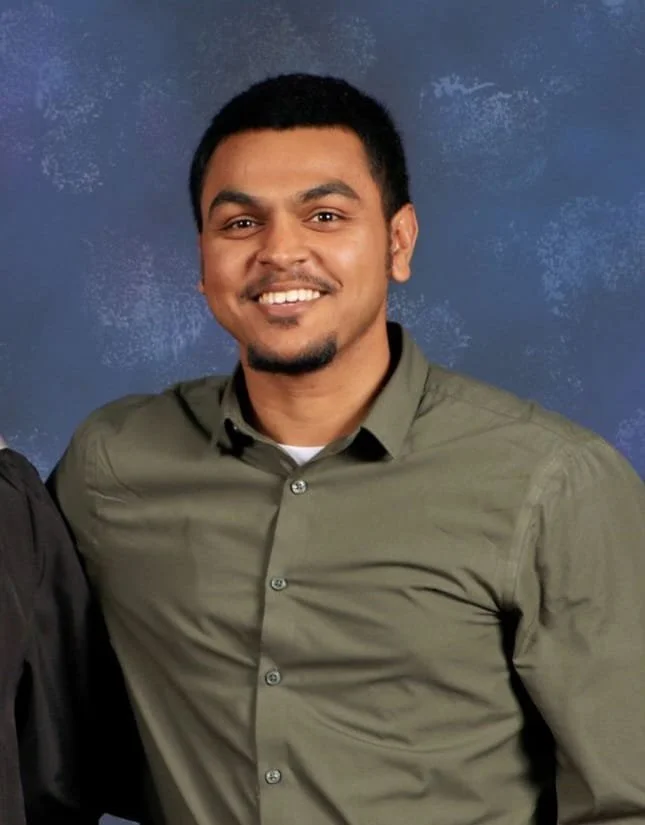 A young man with dark hair, light brown skin, wearing a black sweater, is taking a selfie against a plain light-colored wall.