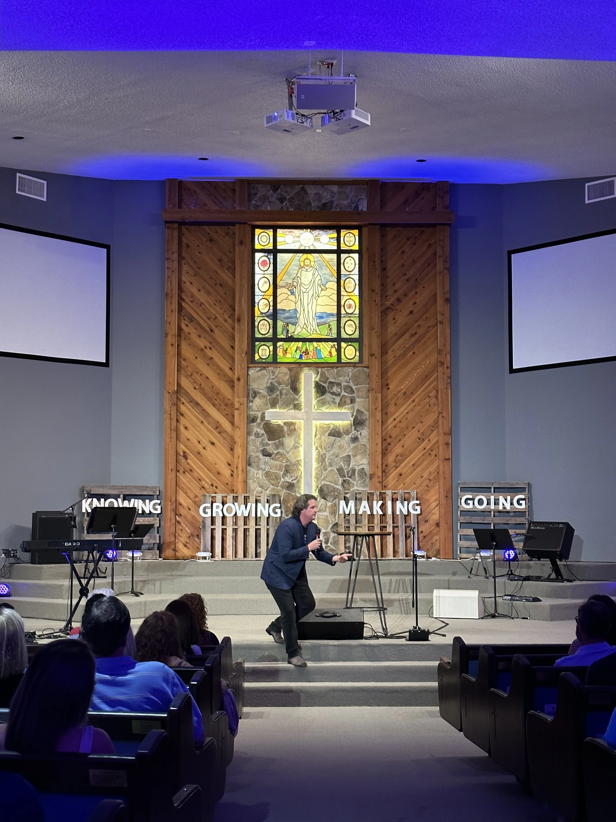A man speaking on stage in a church setting with a stained-glass window, a cross, and signs that read 'Knowing,' 'Growing,' 'Making,' and 'Going,' while an audience listens.
