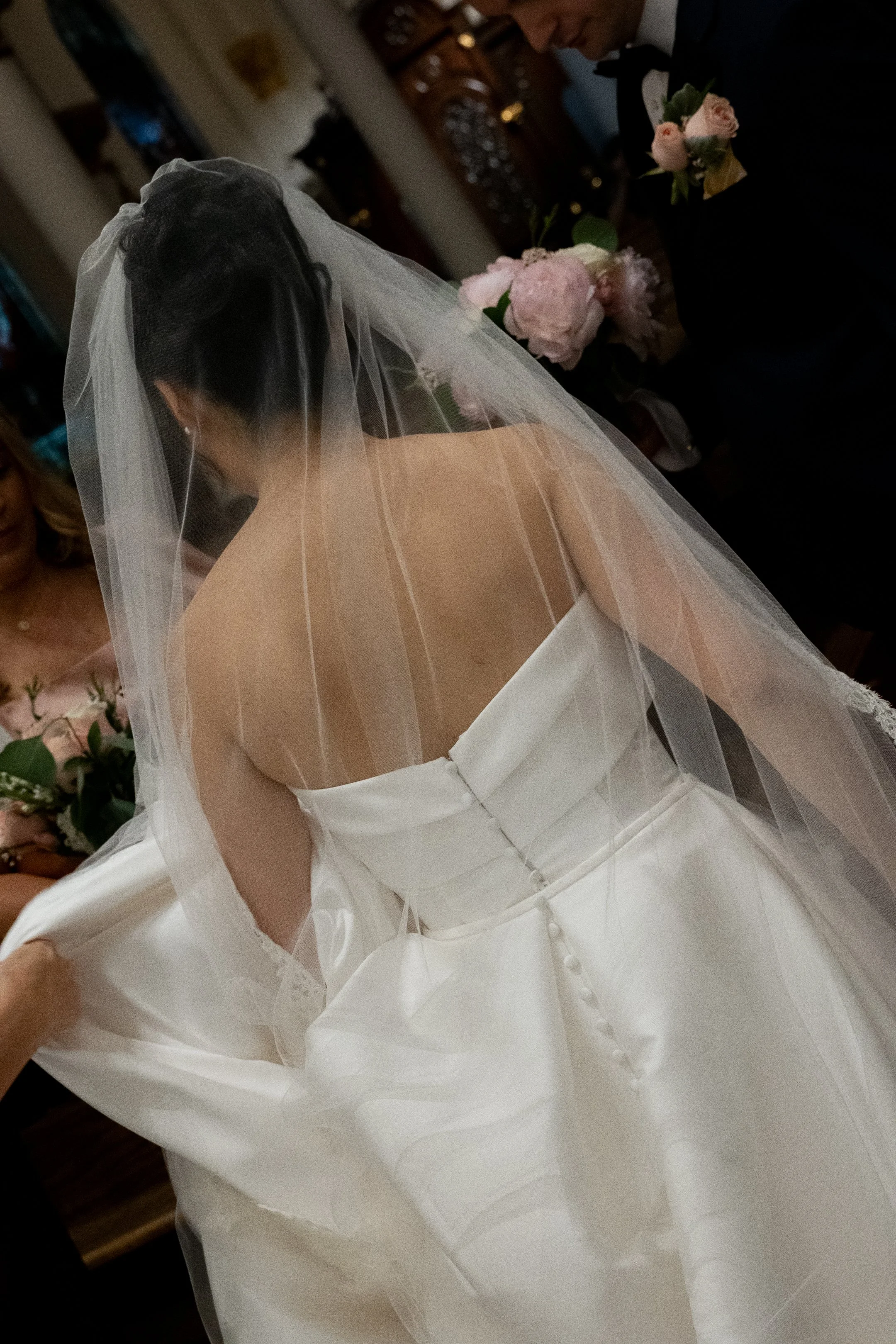 Bride in a strapless white wedding gown with a long veil, surrounded by people, holding a pink bouquet.