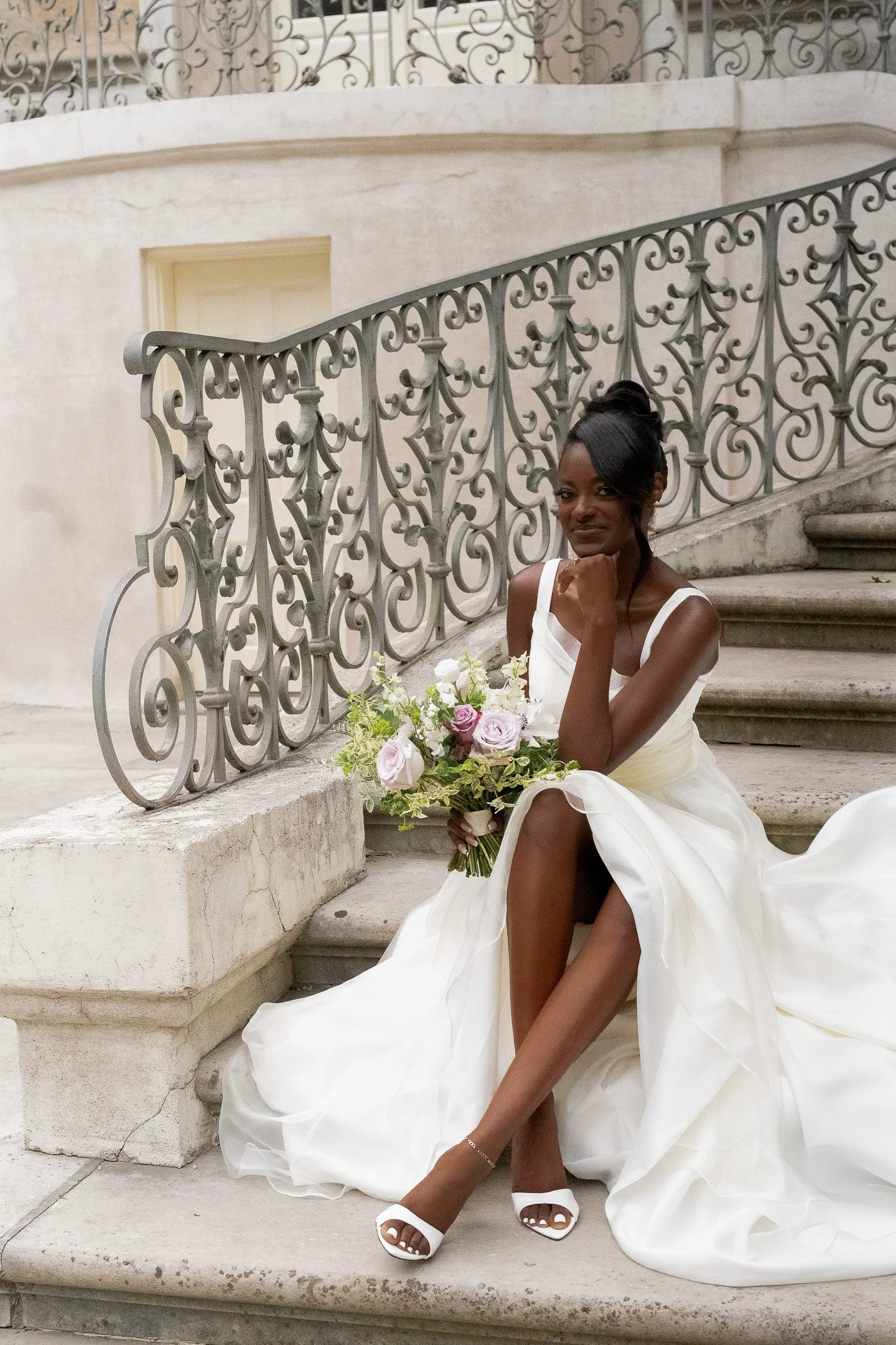 Bride sitting on stairs with bouquet, wearing a white dress and sandals, next to ornate railing.