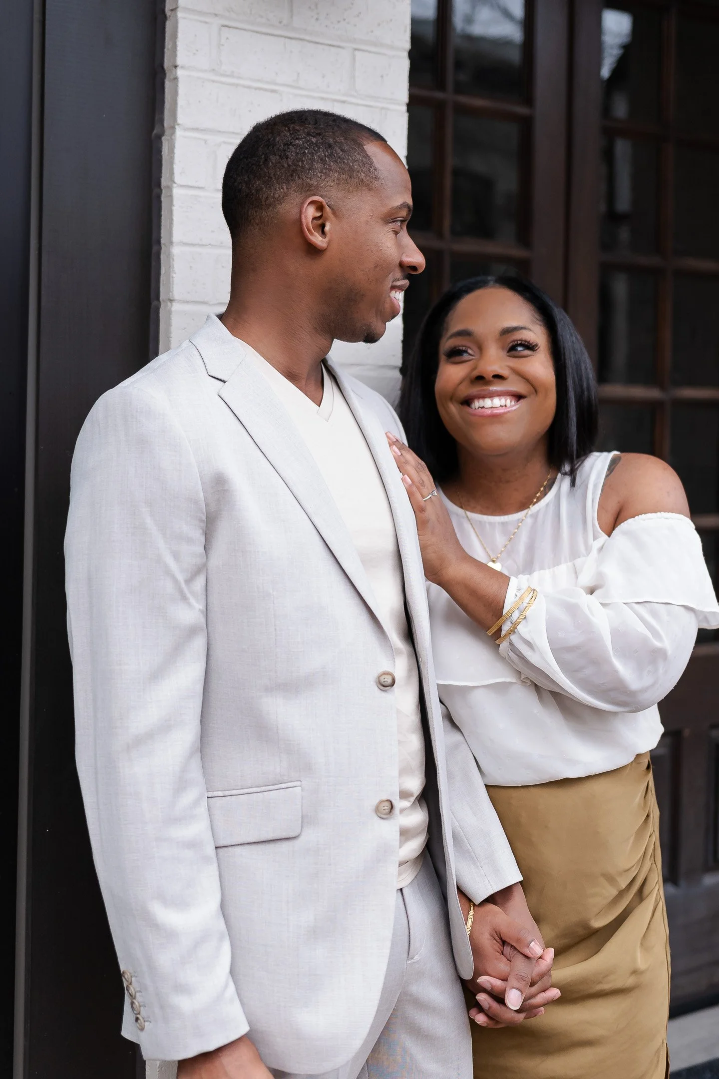 A couple standing closely, dressed stylishly. The man is in a light gray suit, and the woman is in a white off-shoulder top and a tan skirt. They are smiling and holding hands in front of a building with large windows.