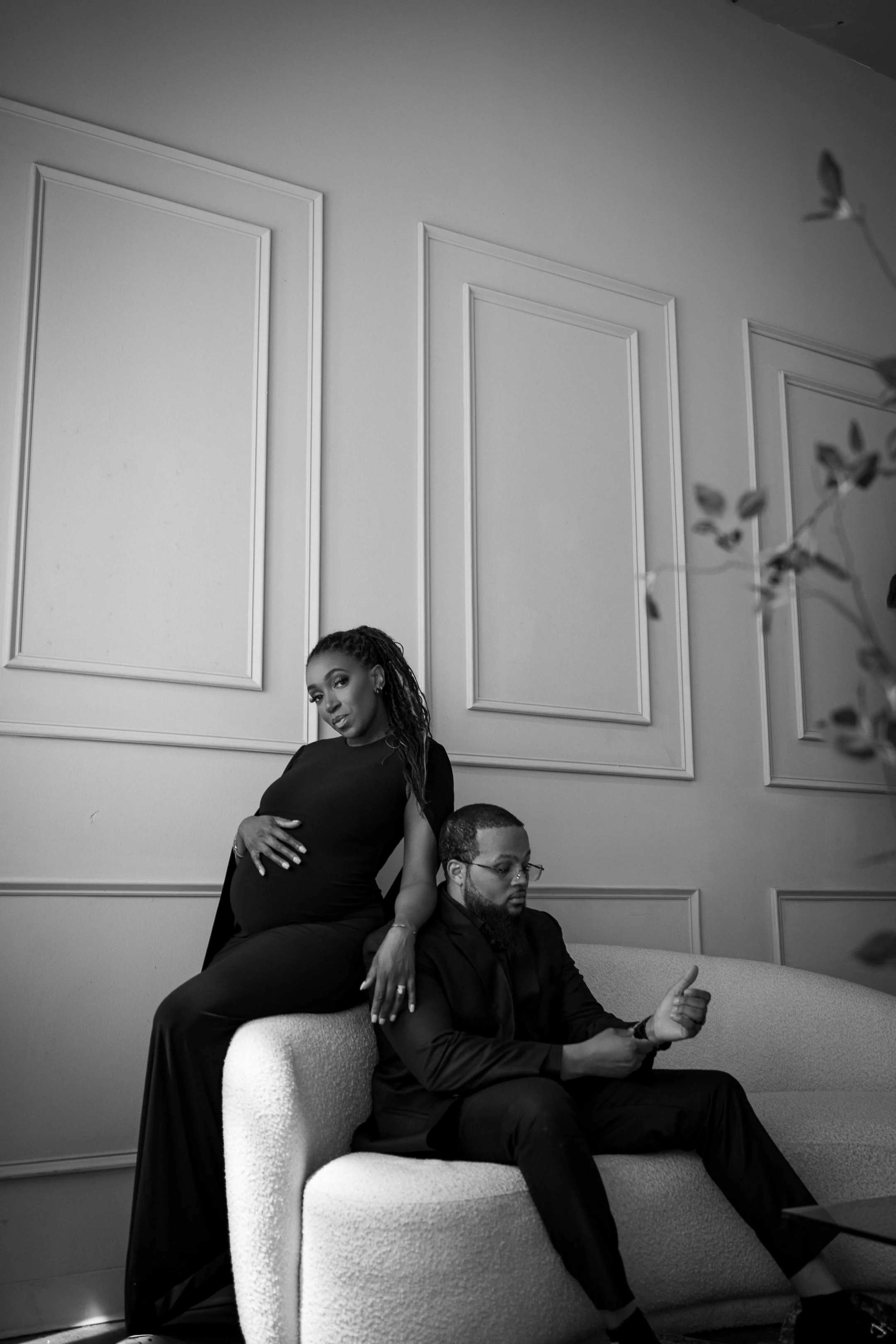 A pregnant woman and a man sitting on a couch in a room with decorative wall panels. The woman is standing, resting her hand on her stomach, while the man is looking at his phone and giving a thumbs up.