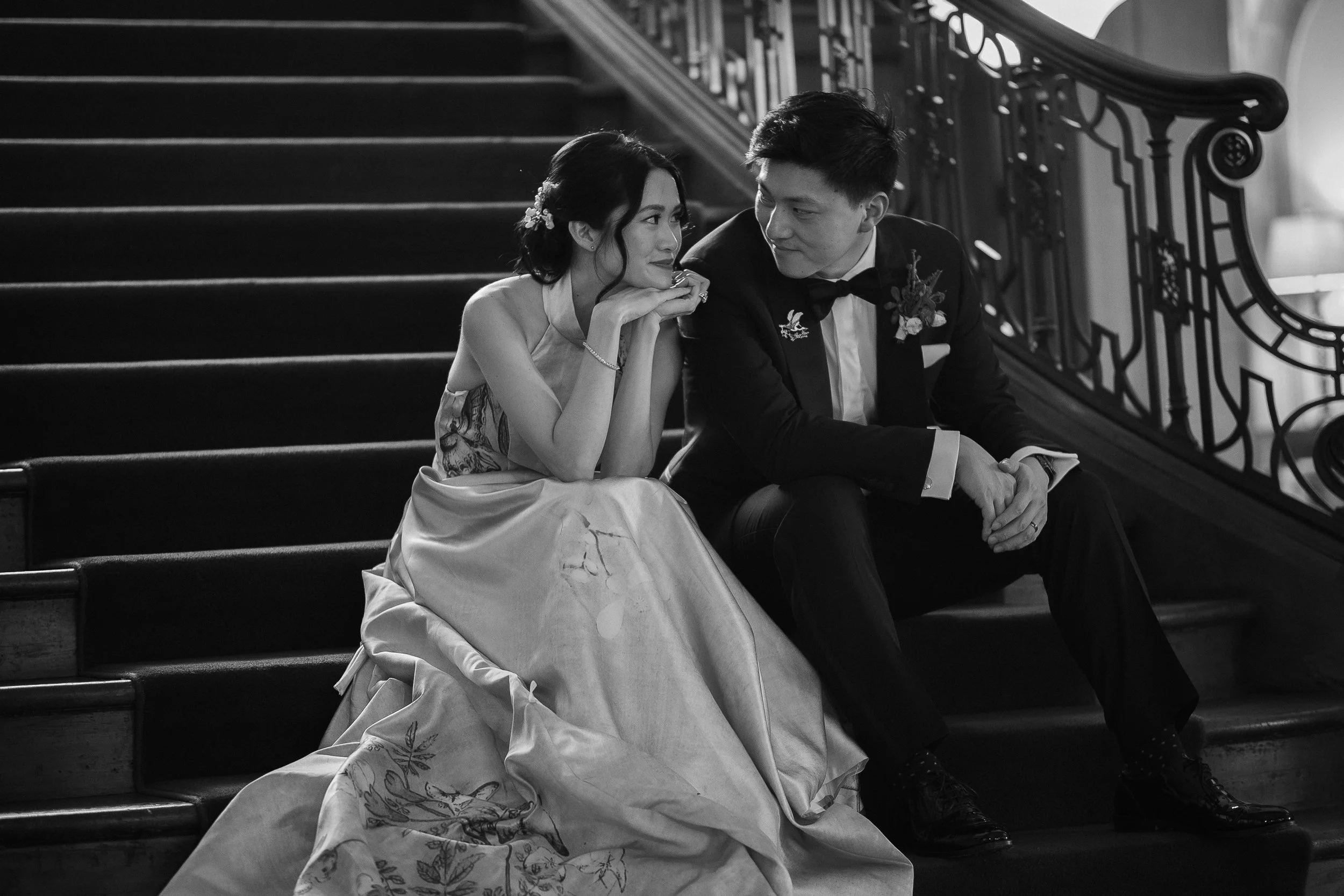 A black-and-white photo of a woman and a man sitting on a staircase, dressed in formal attire, gazing into each other's eyes in an intimate moment.
