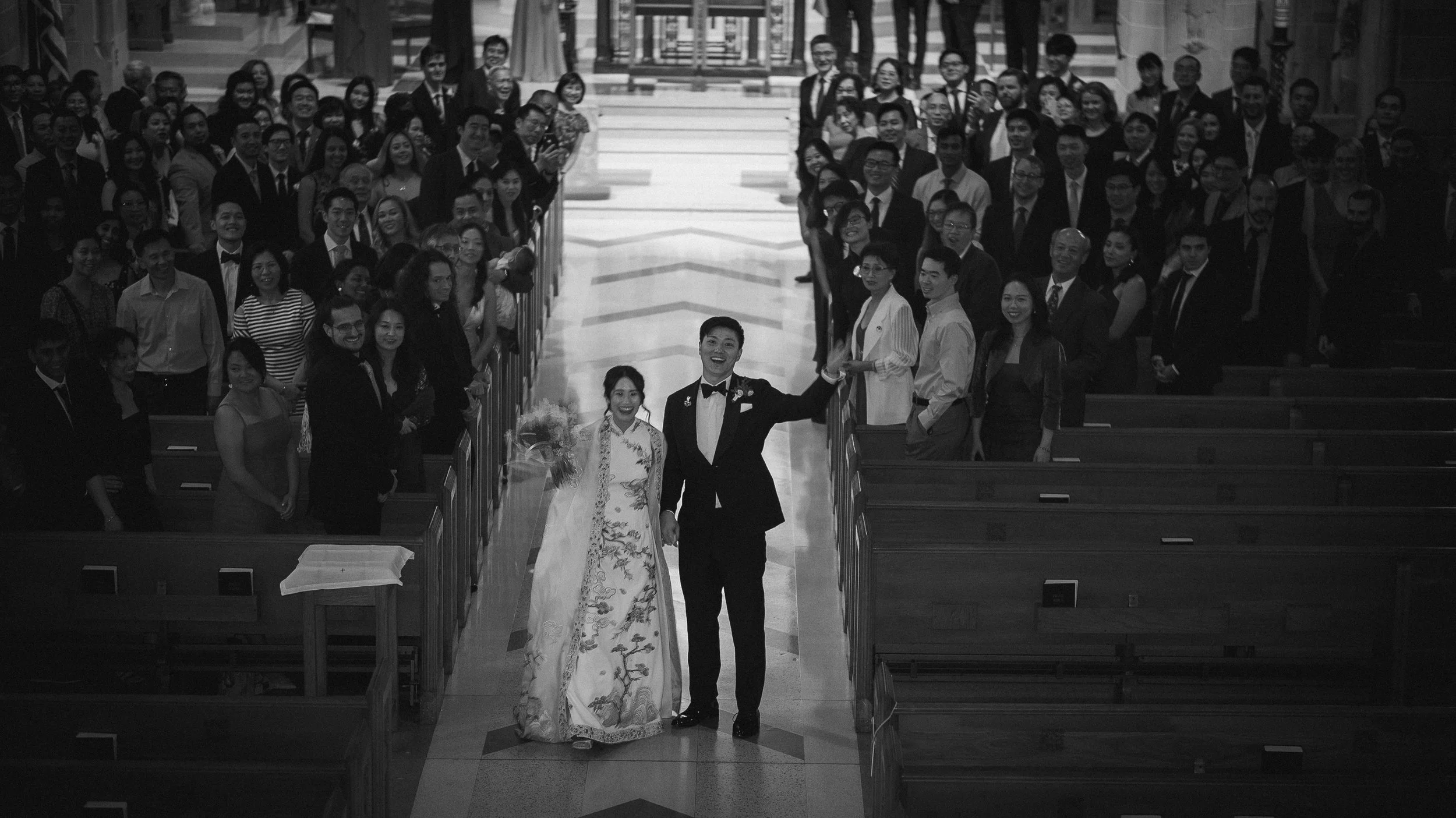 A bride and groom walk down the aisle of a church, holding hands, with family and friends on either side, smiling and clapping.
