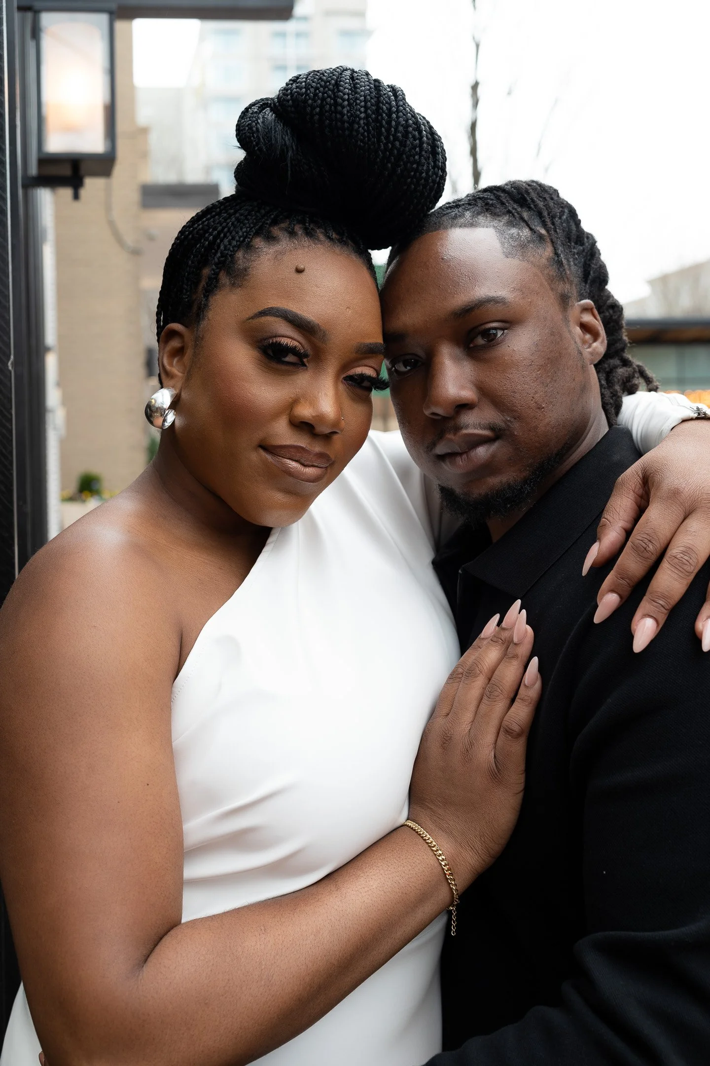 A couple posing closely. The woman is wearing a white dress with a bracelet, and the man is in a black outfit. They are outside, standing against an urban background.