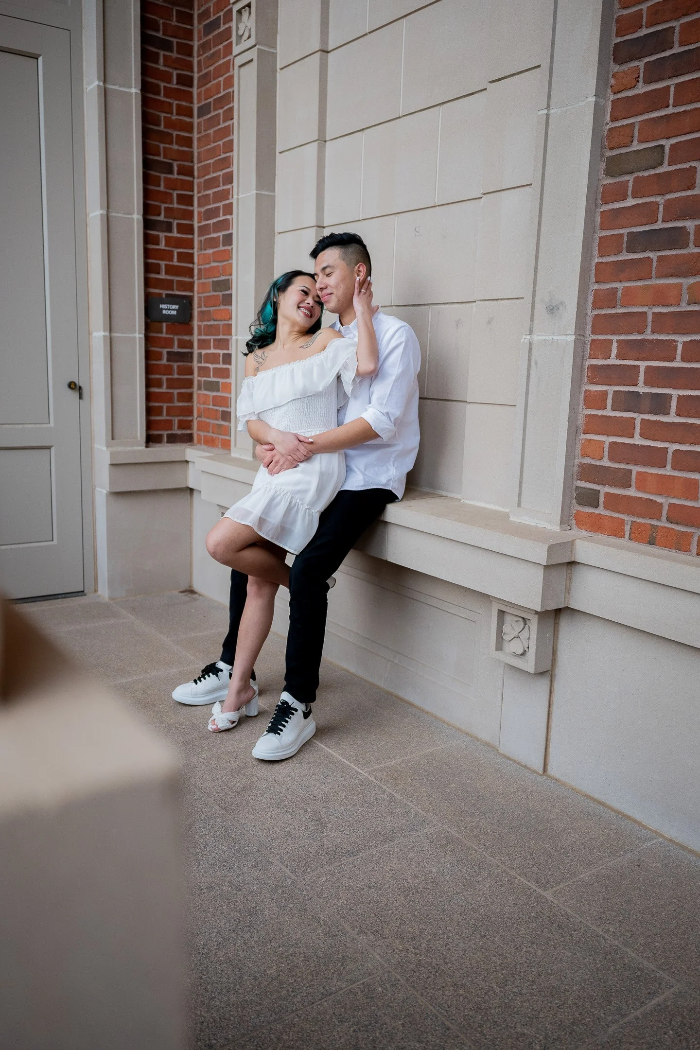A couple sits on a stone ledge in an indoor setting with brick and stone walls. The woman, wearing a white off-the-shoulder dress, sits on the man's lap, who is dressed in a white shirt and black pants. They appear affectionate, smiling and embracing