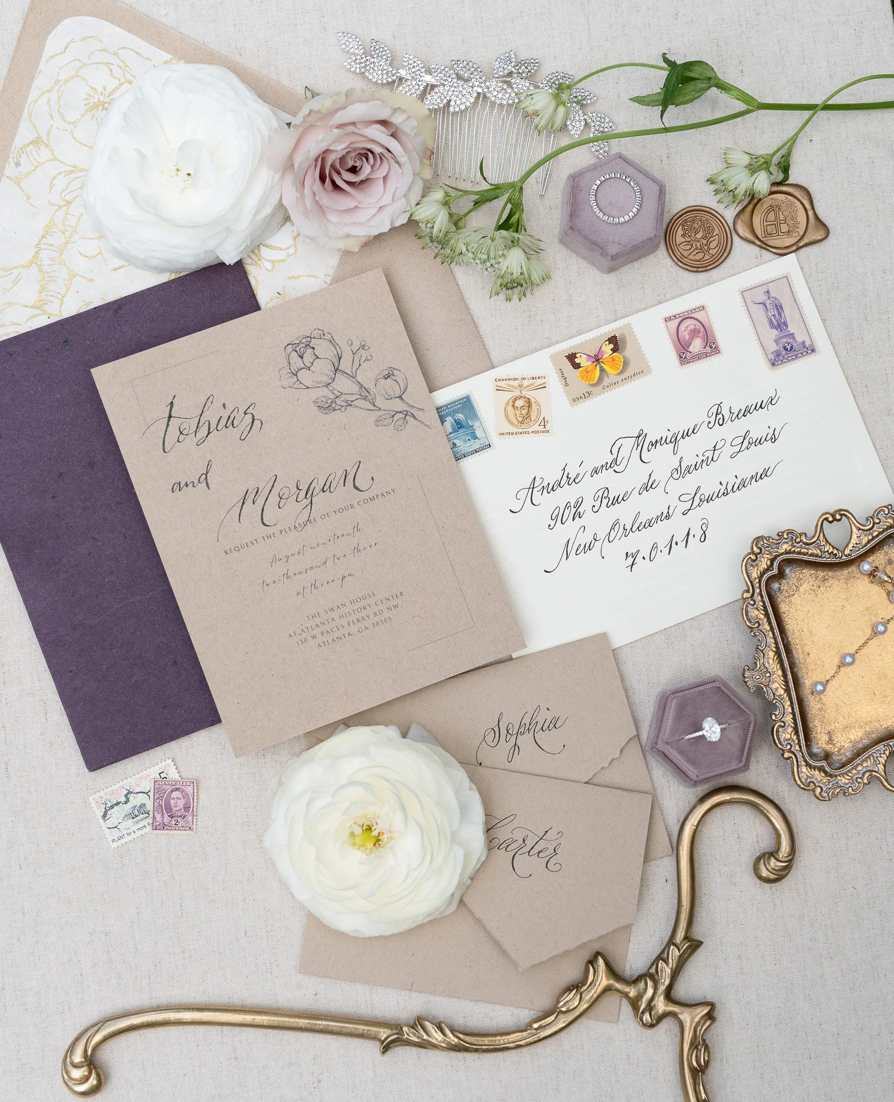 Vintage-themed wedding invitation set with floral and calligraphy elements, featuring a rose, white flower, purple and brown envelopes, stamps, wax seals, a diamond ring in a box, and a gold frame, displayed against a textured background.