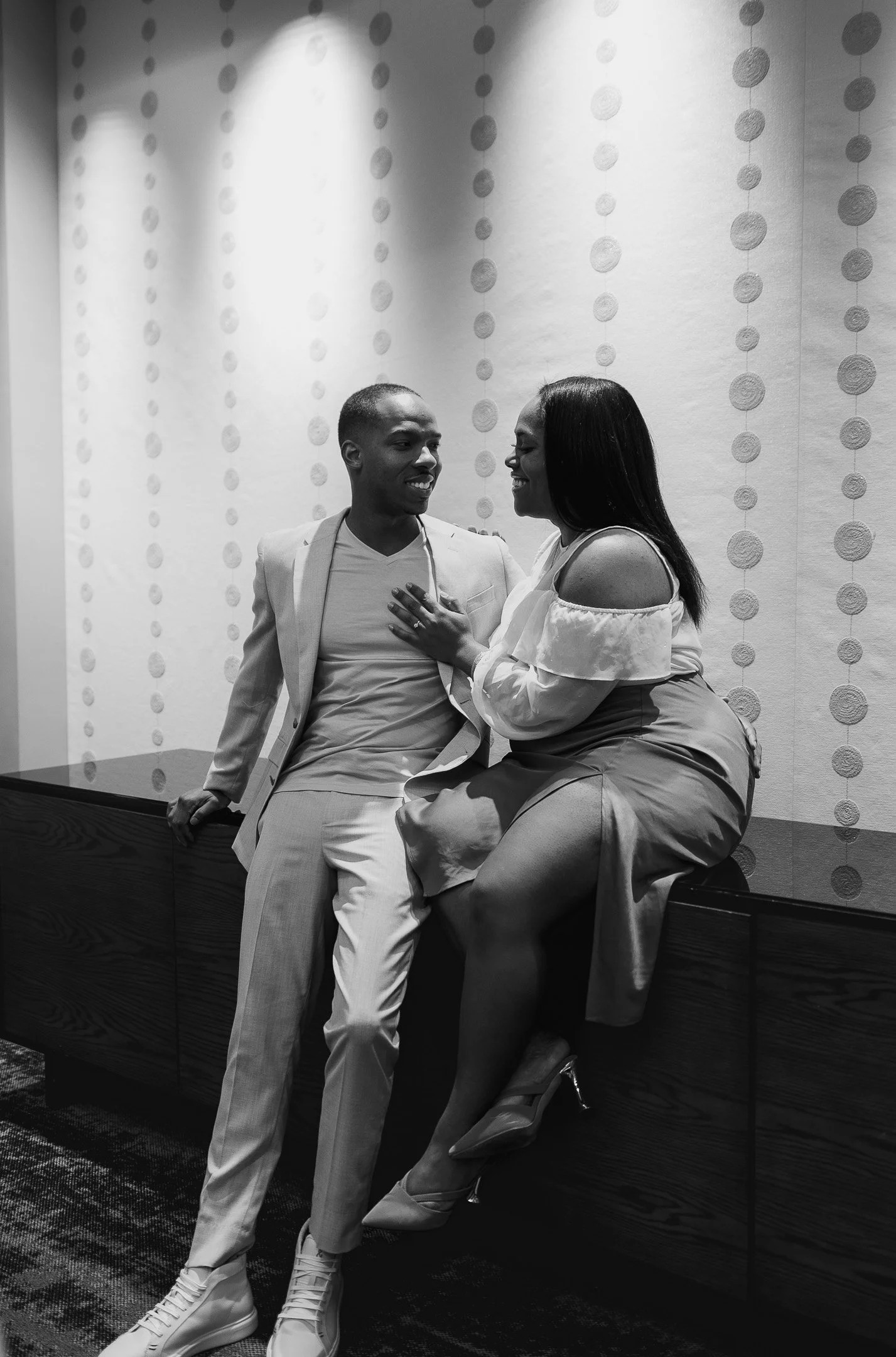 A black and white photo of a couple smiling and engaged in conversation. The man is wearing a suit and leaning against a cabinet, while the woman, in a dress, is sitting on the cabinet, smiling at him.
