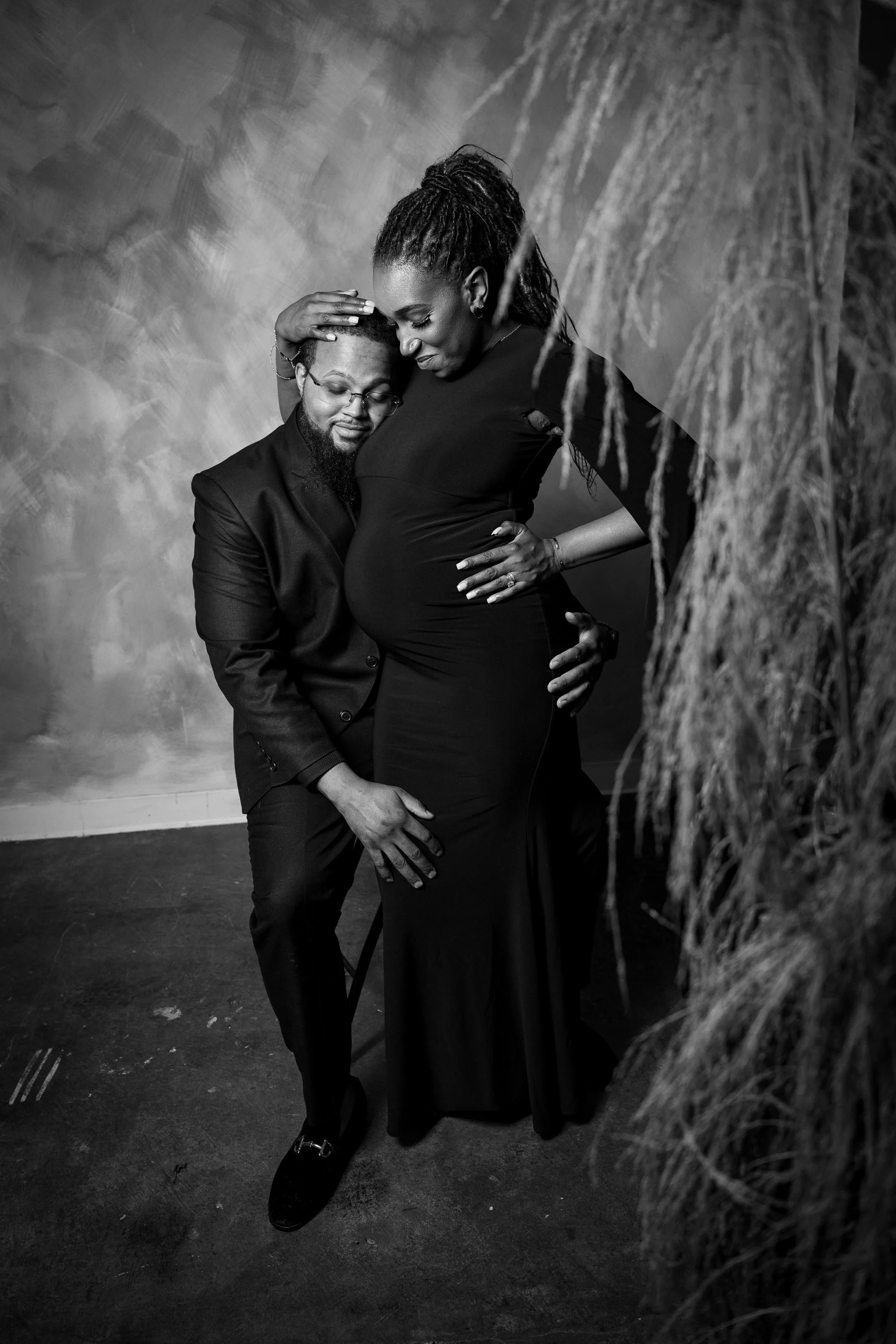 A pregnant woman and a man, likely her partner, share an intimate moment. The man is kneeling and resting his head against her belly, while she gently rests her hand on his head and her other hand on her stomach. The scene is in black and white, with