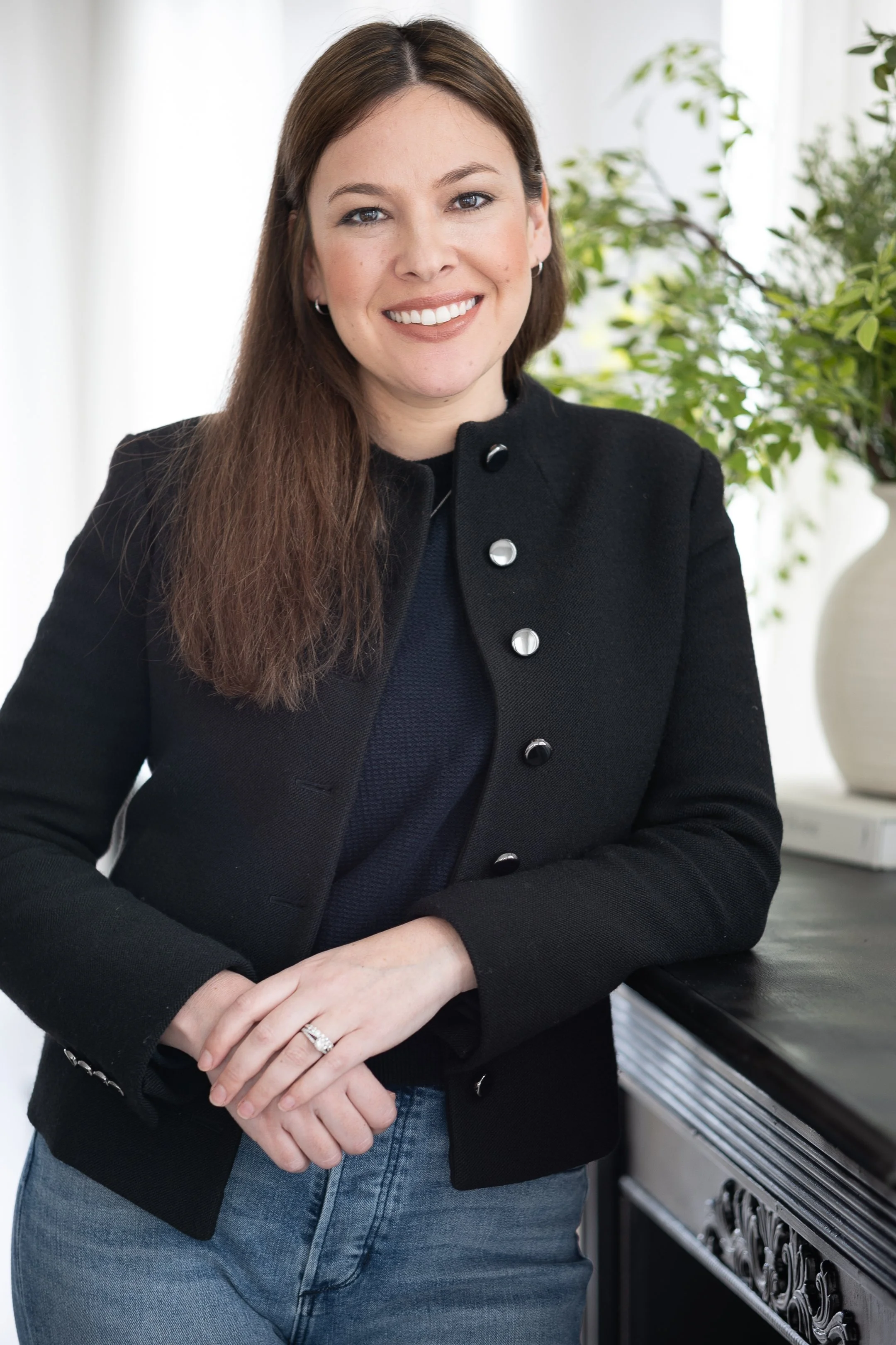 Smiling woman in black jacket and jeans with long brown hair standing by a table with greenery in the background.