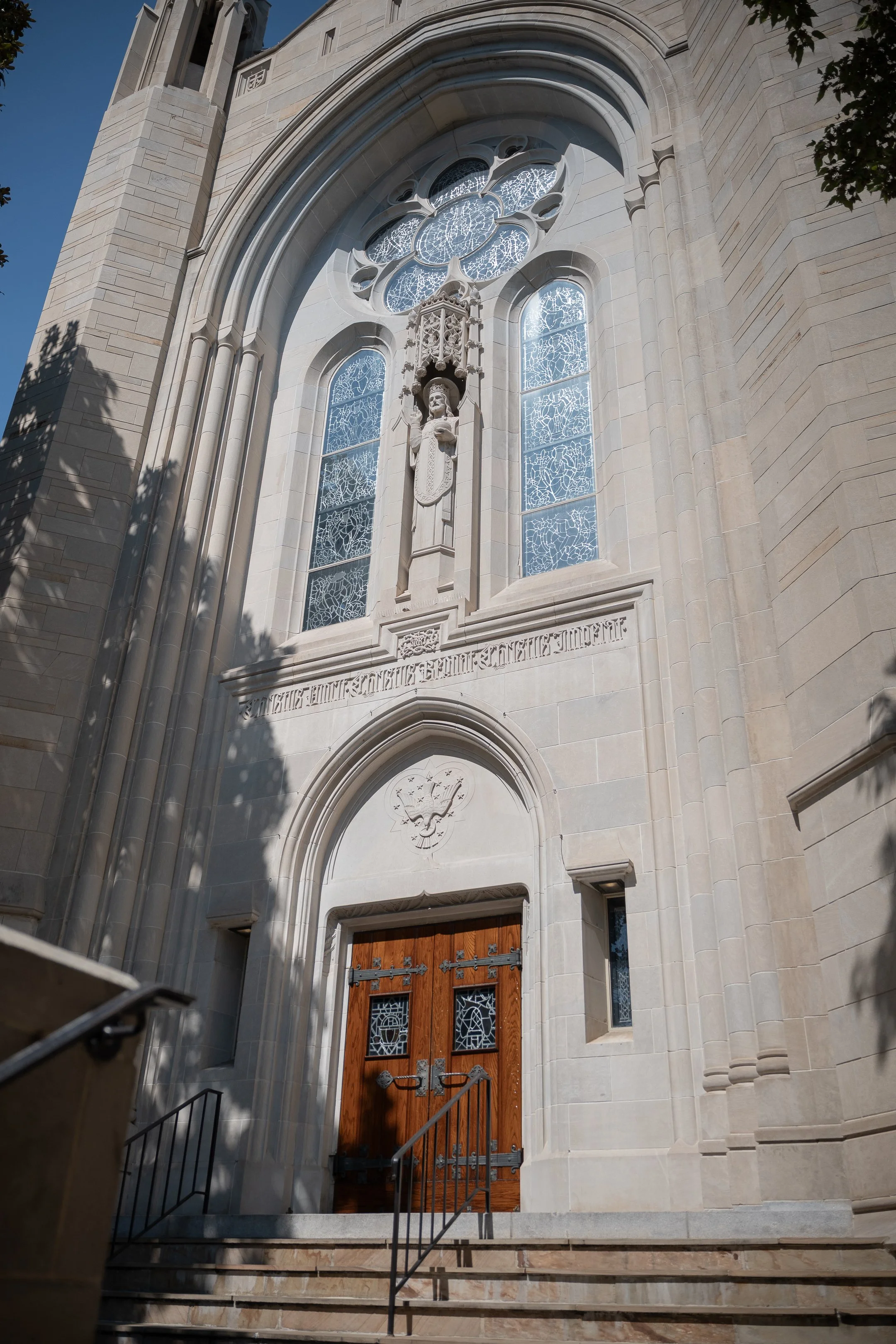 View of a church's entrance with a wooden door, stone facade, stained glass windows, and a statue of a saint above the door.