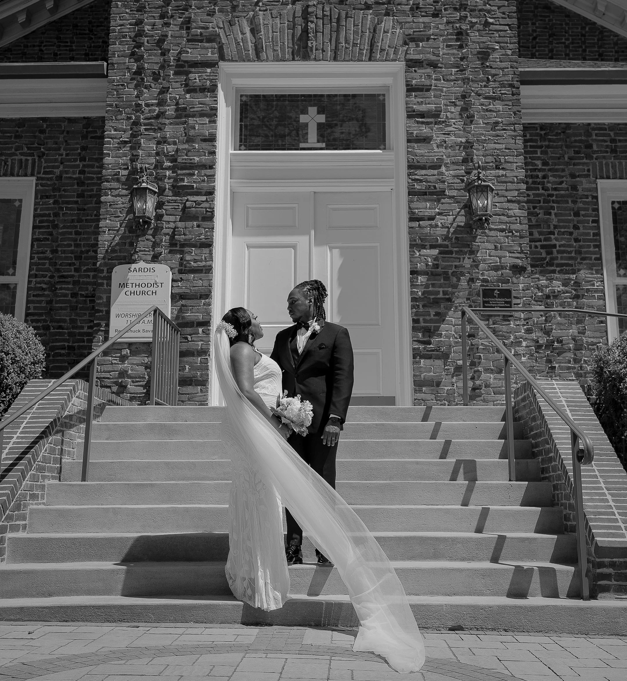 Bride and groom standing on the steps of Sardis Methodist Church, sharing a loving gaze. The bride holds a bouquet and wears a veil, while the groom is in a suit. Black and white photography emphasizes the romantic setting.