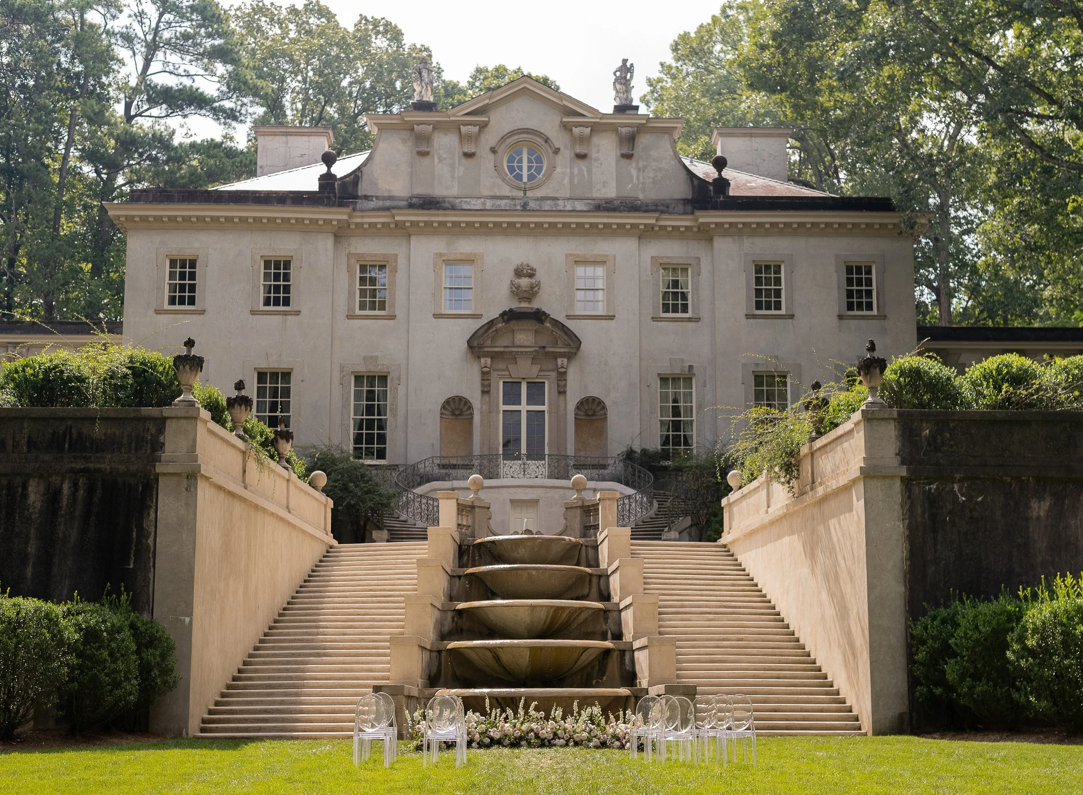 Front view of a large historic mansion with a formal garden, featuring stone steps, fountains, and decorative statues, surrounded by trees.