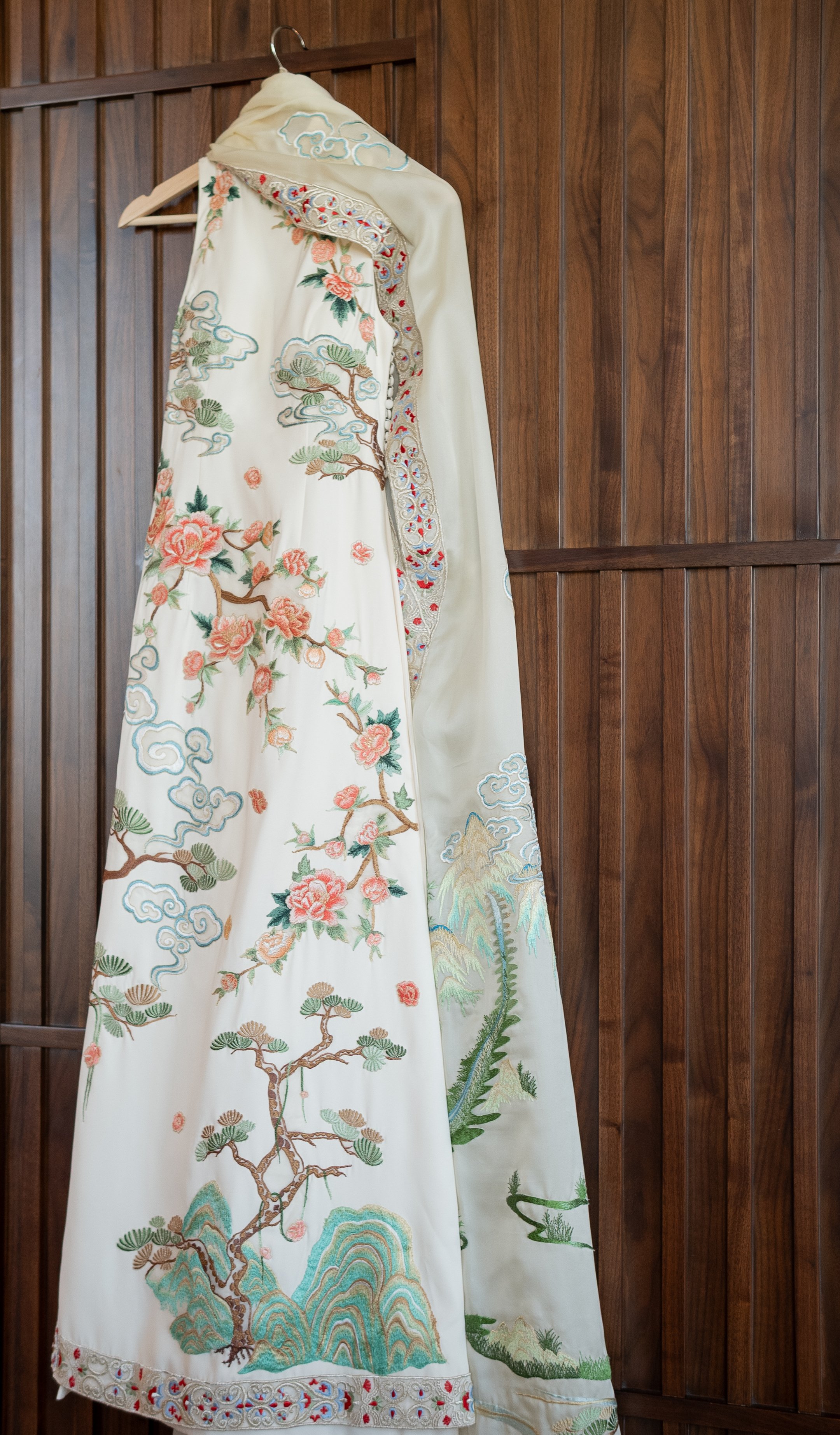 An embroidered white robe with colorful floral and nature designs hanging on a wooden wall.