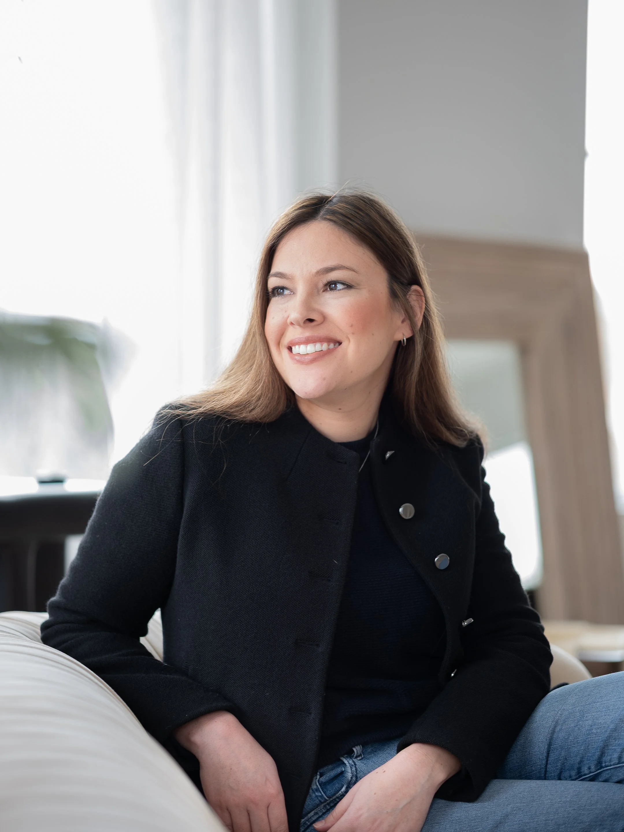 Woman smiling, wearing a black jacket, sitting on a white sofa, with a mirror in the background.
