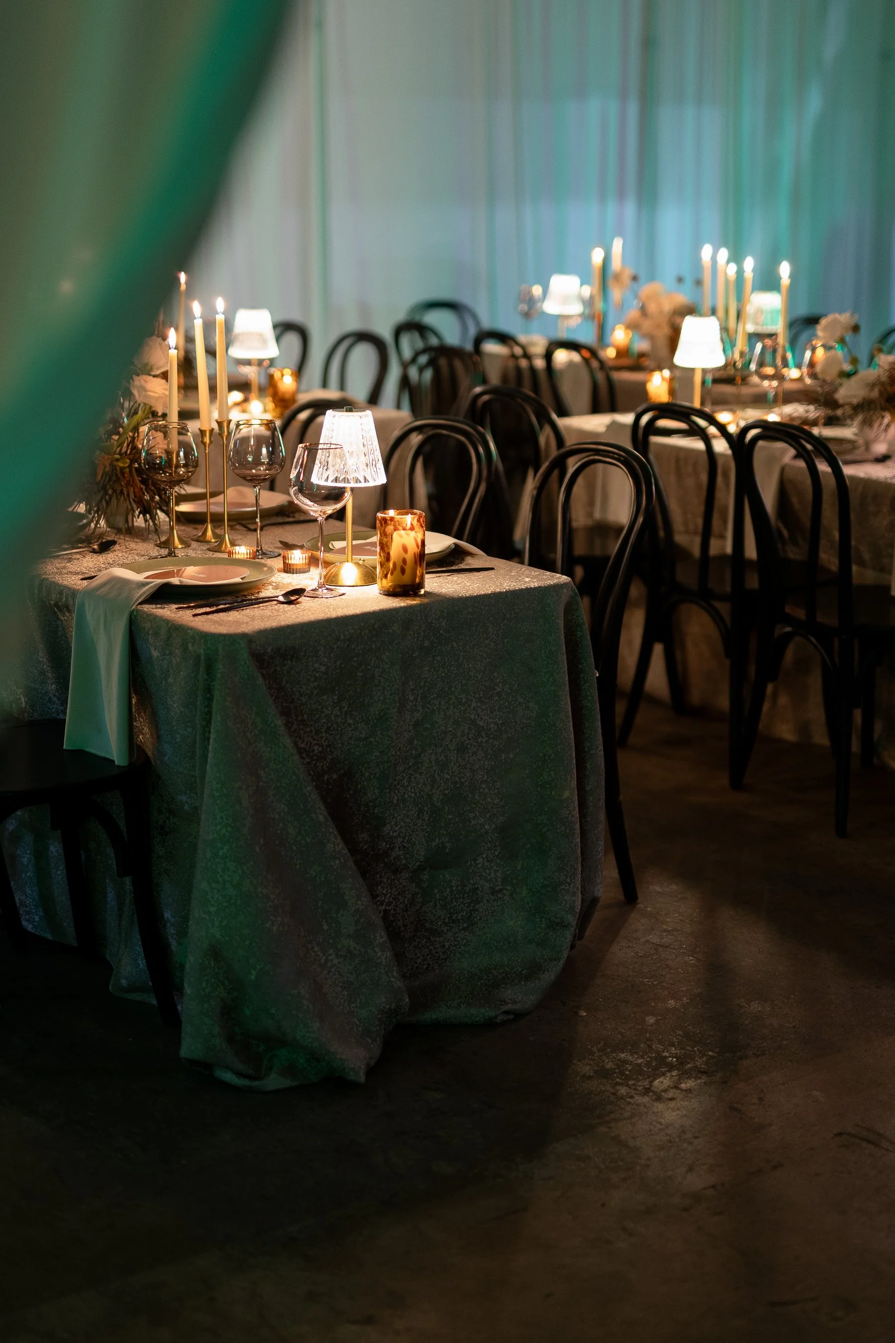 Elegant dining setup with tables covered in textured tablecloths, set with plates, wine glasses, candlesticks, and decorative lamps. Dim lighting and black chairs create an intimate atmosphere.