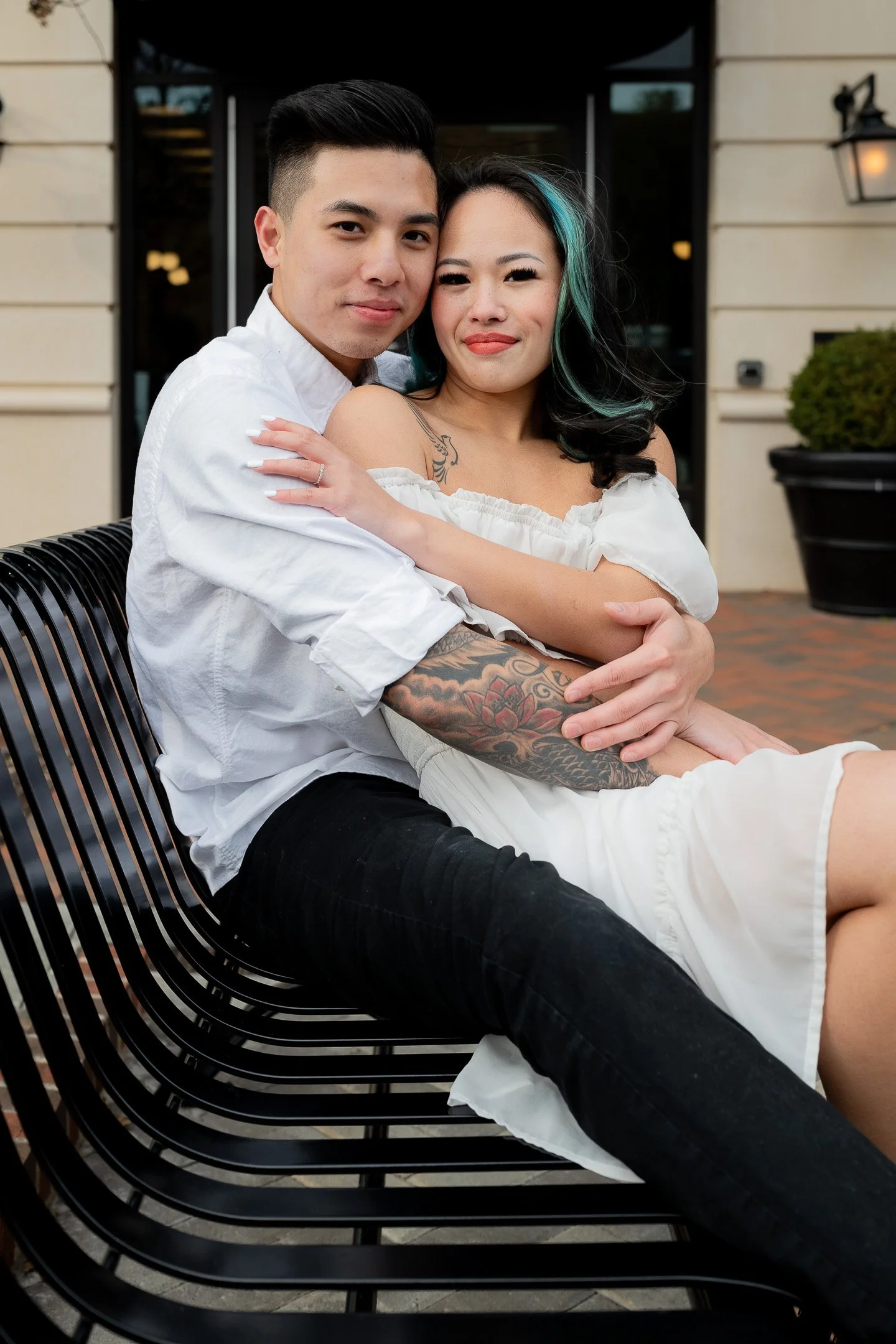 A couple sitting on a bench, with the woman wearing an off-shoulder white dress and the man in a white shirt. The woman has tattoos on her arm, and her hair has green streaks.