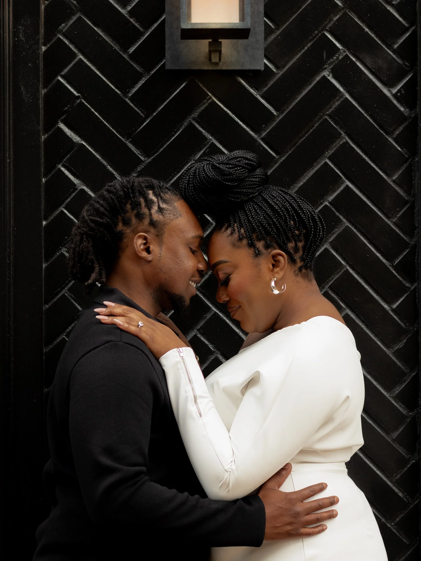 A couple embracing in front of a black herringbone patterned wall, highlighting the woman's engagement ring.