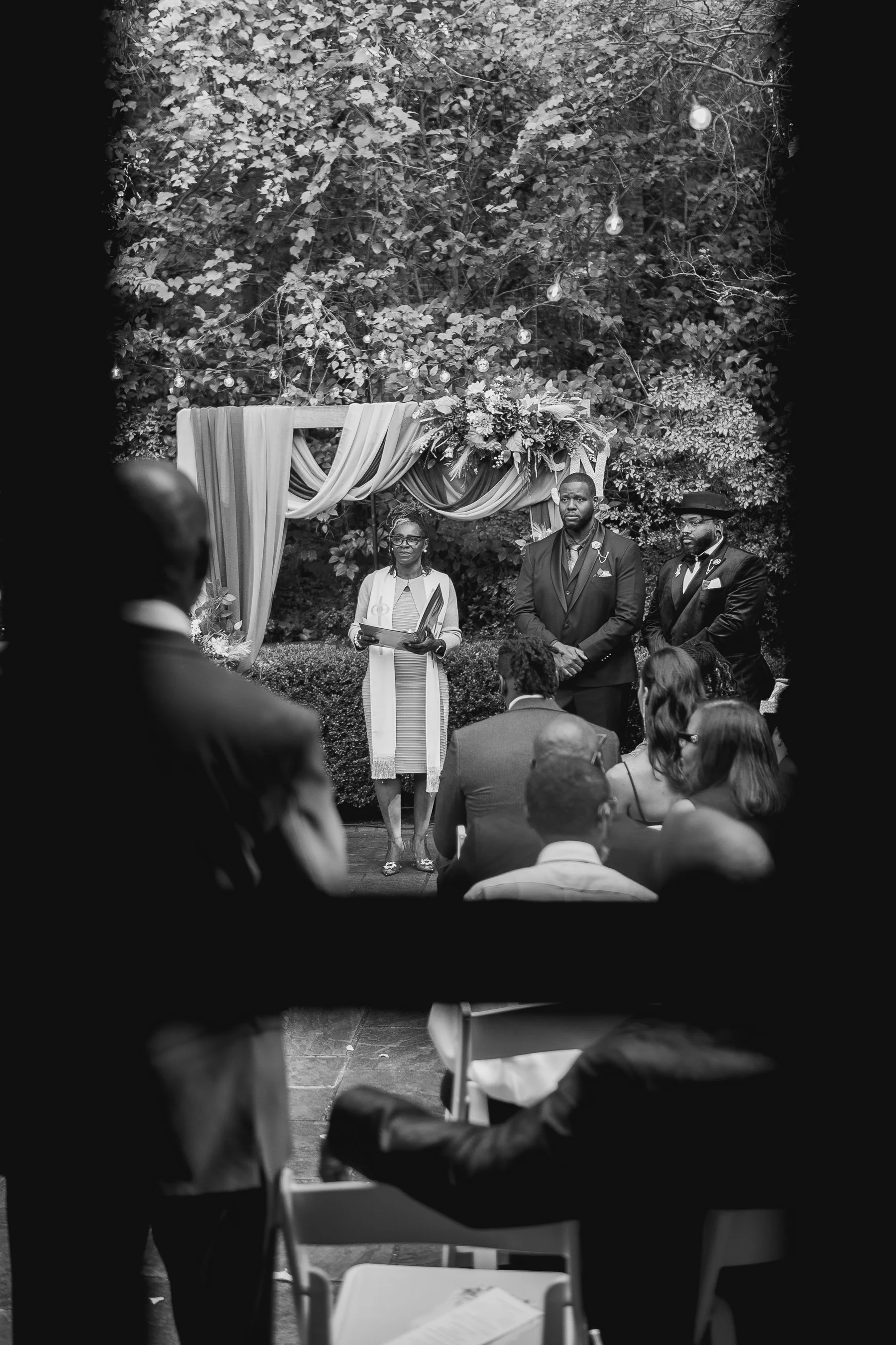 Black and white photo of an outdoor wedding ceremony with an officiant and two groomsmen under a floral arch, guests seated and watching.