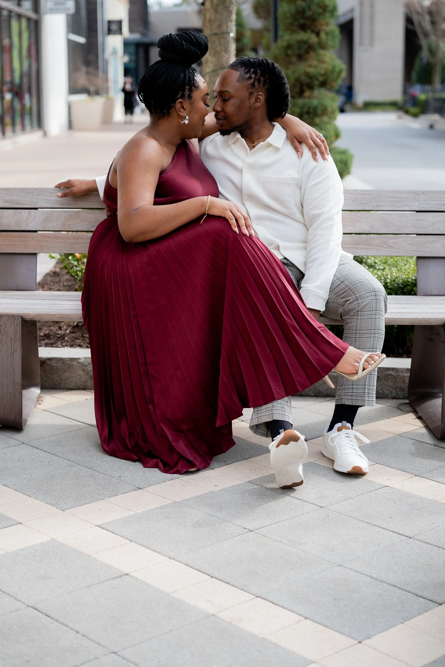 Couple sitting on a bench outdoors, woman in burgundy dress and man in white shirt and plaid pants, embracing and looking at each other.