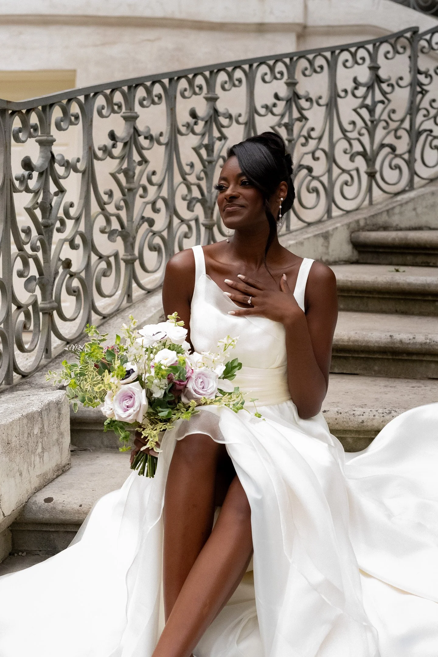 Bride in white wedding gown holding bouquet, sitting on stone steps with ornate railing.